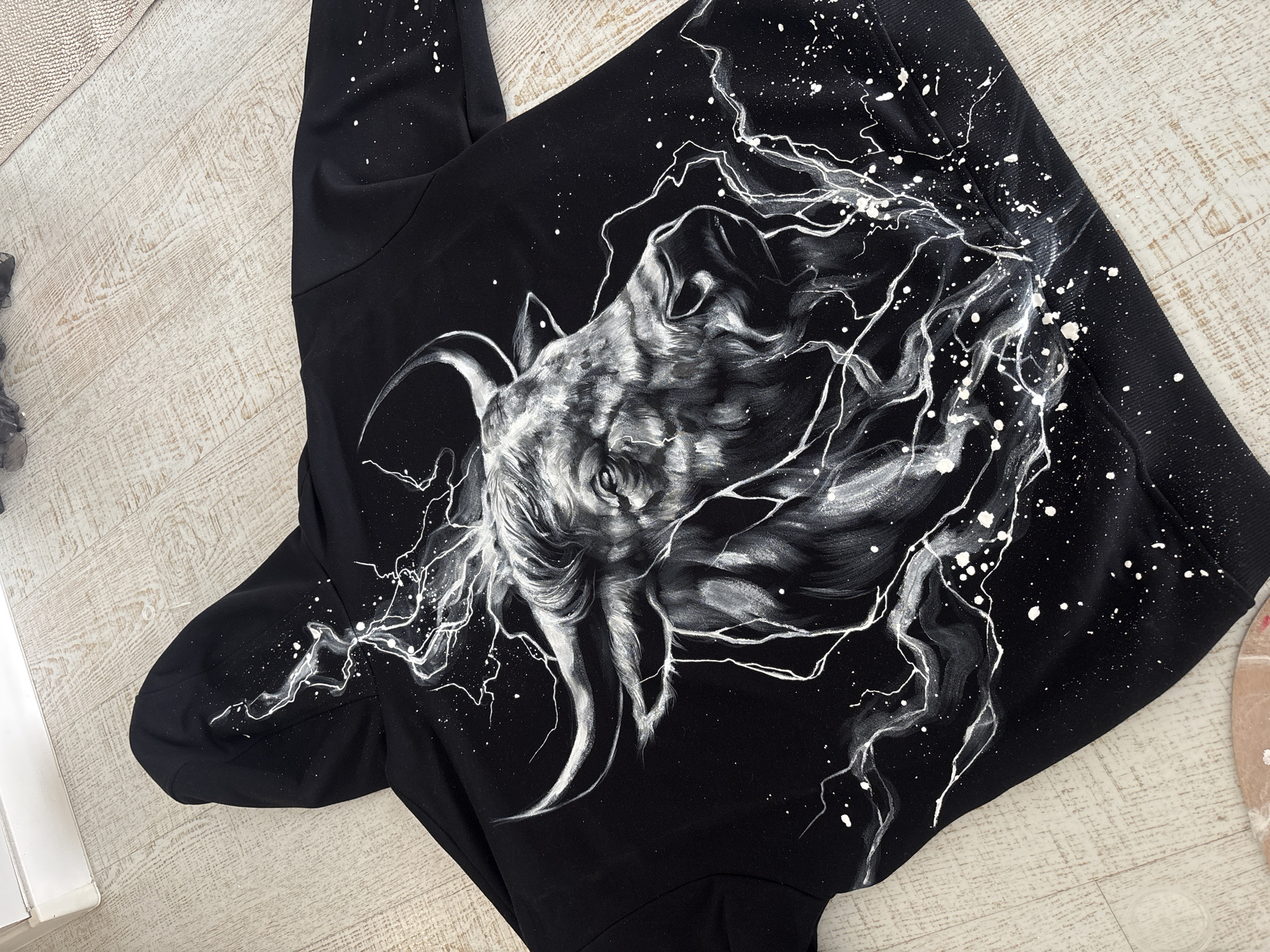A black garment, likely a hoodie or sweatshirt, is laid flat on a light-colored wooden floor with distinct grain patterns. A textured, light beige rug is partially visible in the top left corner of the frame.
The central subject is an elaborate hand-painted design on the back of the black garment. The artwork features a stylized, powerful-looking animal head, resembling a goat or ram, rendered in shades of white, grey, and black. The animal's head is depicted with prominent, curved horns and an intense, almost ferocious expression. White, lightning-like streaks emanate from around the animal's head, suggesting energy or motion. The design is further enhanced by numerous white paint splatters scattered across the black fabric, creating a dynamic, energetic, and slightly distressed aesthetic.
The garment's fabric appears soft and slightly wrinkled in places, indicating its material. The sleeves and lower hem of the garment are visible, showing the continuity of the black fabric.
No specific activity is taking place beyond the display of this unique garment. The indoor setting provides no cues about the time of day or weather. There is no visible text in the image. A small, light brown, circular object, possibly a painter's palette or a coaster, is glimpsed on the right edge. The overall impression is a showcase of custom wearable art.