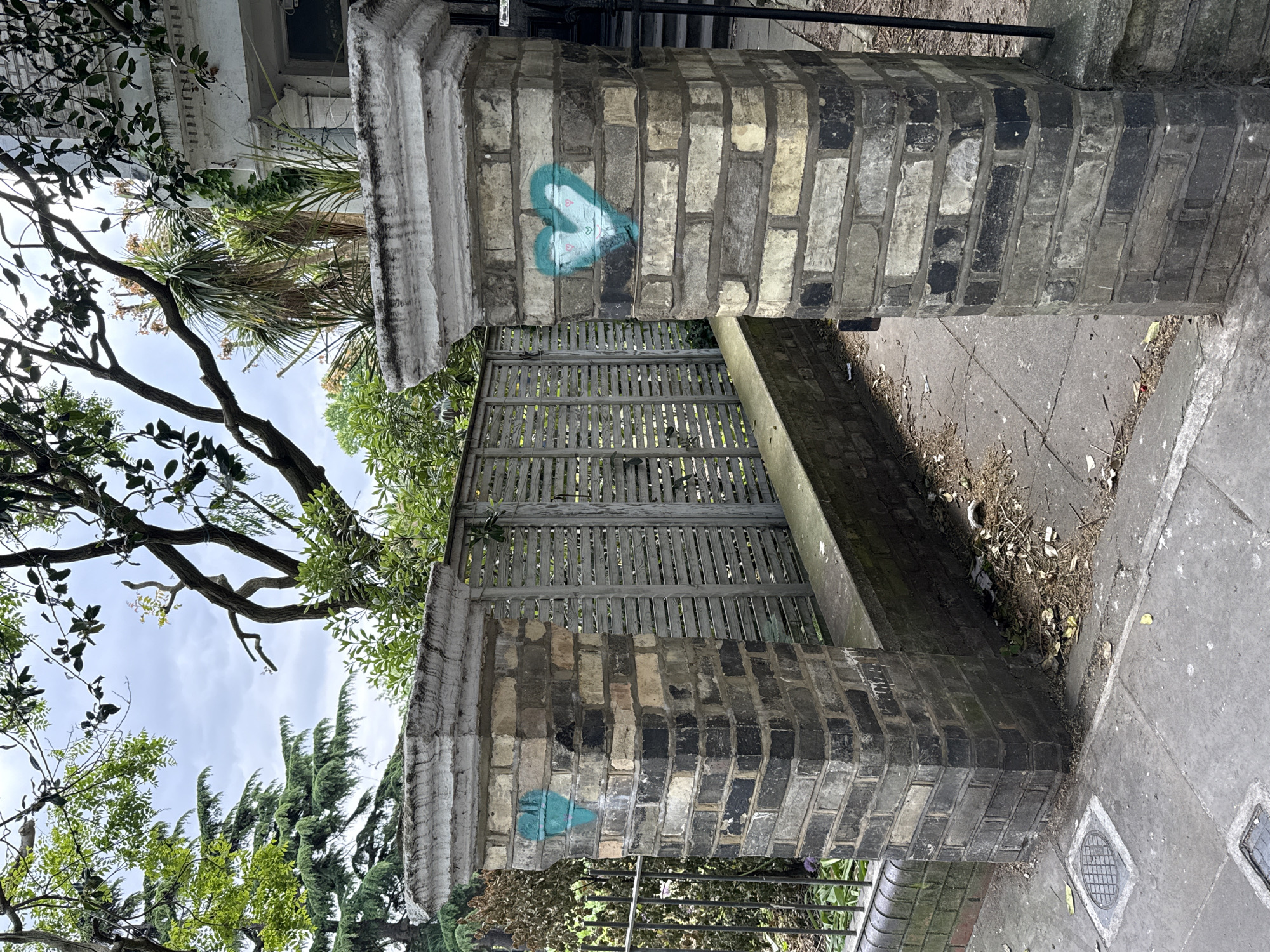 The image shows a brick gatepost in an urban environment. The gatepost is made of weathered bricks, some darker than others, and topped with a decorative concrete cap. There is blue heart-shaped graffiti on the brick surface. Behind the gatepost is a wooden fence with horizontal slats. Trees and foliage are visible in the background. A section of pavement and a drain cover are visible on the right side of the image. It is not possible to definitively determine the exact location in London from this image.