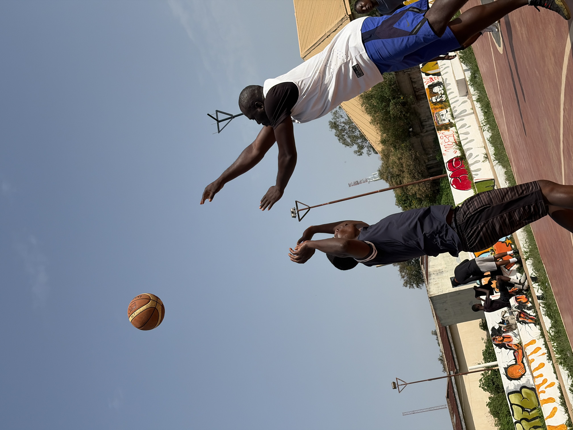 Two men are playing basketball on an outdoor court. One man is in mid-air, reaching for a basketball floating in the air. The other man is jumping to try and block the first man from catching the ball. The court has graffiti on the walls. The sky is blue and the court appears to be in Jos, Nigeria.