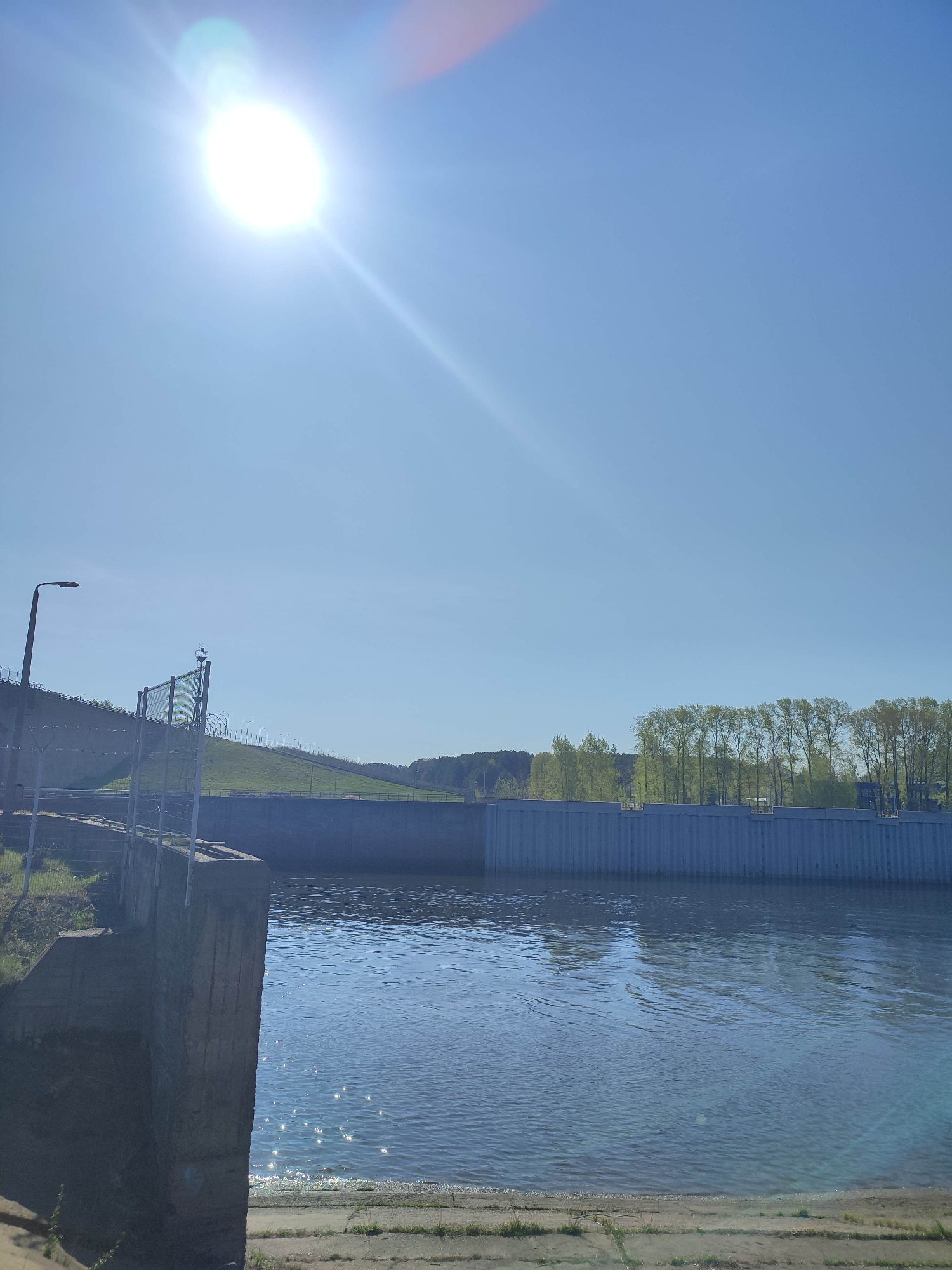 The image depicts a sunny scene with a body of water, likely a river or reservoir, in the foreground. A concrete dam or wall runs across the middle ground, with trees visible beyond it. To the left, a fenced-off area with a concrete structure extends into the water. The sky is clear and blue, with the sun positioned high. The reflections of light on the water create a shimmering effect. There is also a series of light posts along the hill in the background.
Location: Unknown, Unknown.