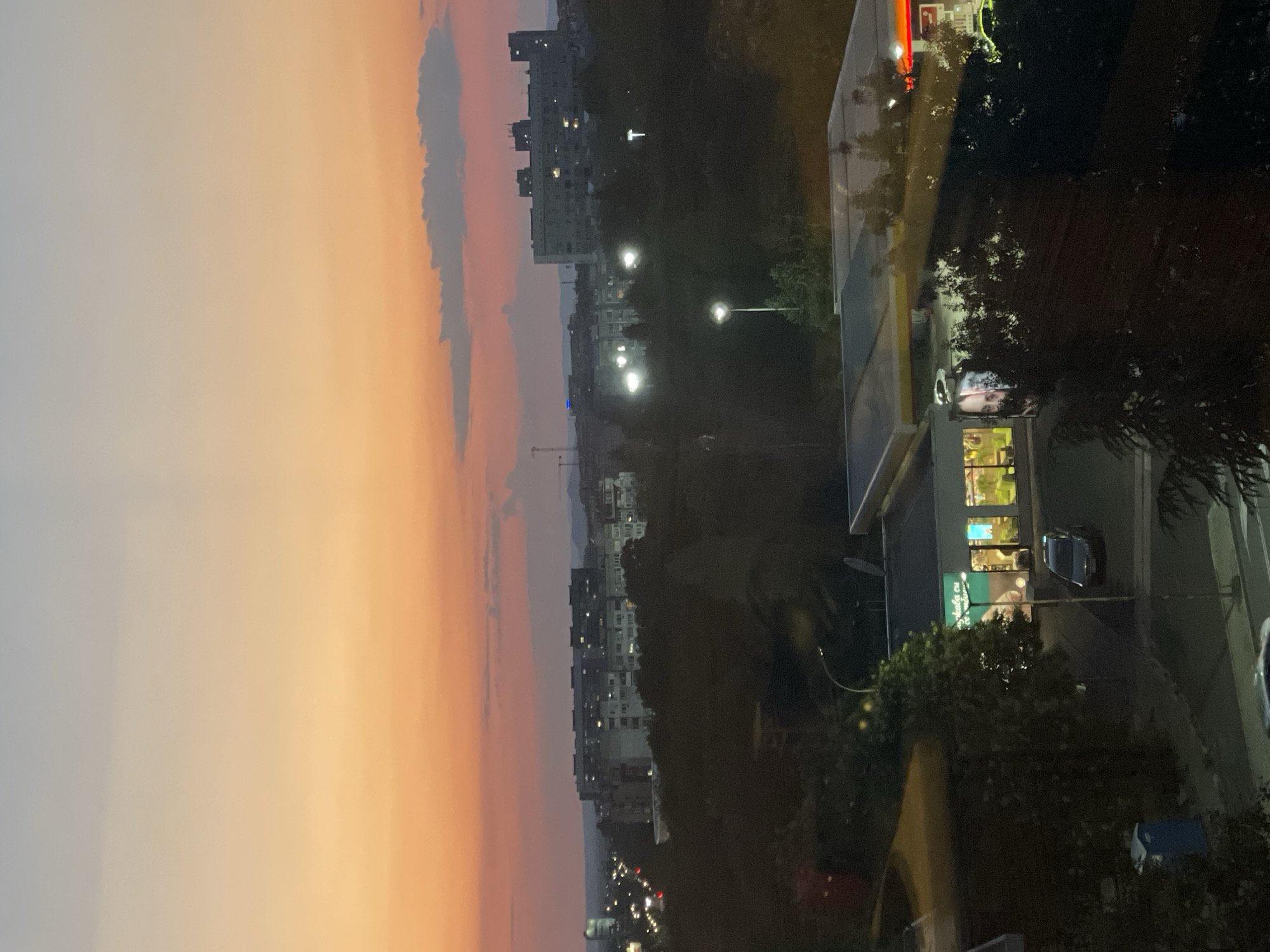 This is a medium-shot, vertical, eye-level photograph of a city skyline in Burgas, Bulgaria at dusk. The sky transitions from a pale blue at the top to a vibrant orange and pink gradient near the horizon, with a single, wispy cloud visible in the upper right quadrant. The middle ground of the image is dominated by dark green trees, obscuring much of the city below. Scattered among the trees are illuminated buildings, appearing as clusters of lights. Prominent in the foreground is a low-rise commercial building with a yellow awning and glass storefronts, revealing the interior. Adjacent to this structure is a gas station with a red and yellow sign. A dark-colored car is parked on the asphalt in front of the commercial building. A blue portable toilet is situated on the left side of the image, near a paved area. The overall atmosphere is that of a city transitioning from day to night, with artificial lights beginning to illuminate the urban landscape against the backdrop of a colorful sunset.