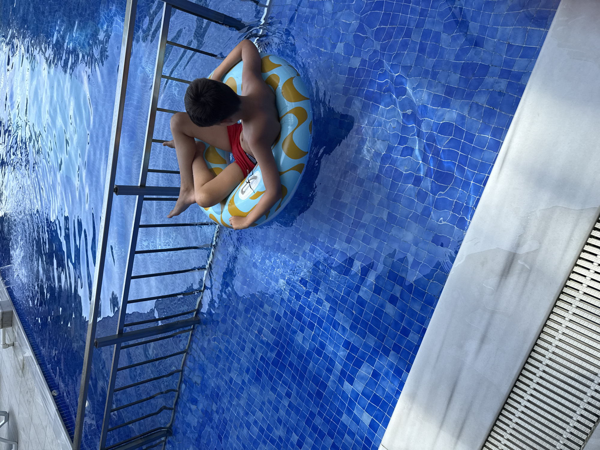 A young boy, seen from behind, rests in a blue and yellow inflatable ring in a tiled swimming pool. He is wearing red swim trunks and his legs are crossed. The pool is surrounded by a metal railing. The image is taken from a high angle, looking down into the pool. The bottom of the pool is visible, with blue tiles creating a grid pattern. The water is clear and rippling. The edge of the pool is a light-colored, solid surface, and next to it is a metal grate drain. The scene appears to be an outdoor pool area, possibly at a hotel or resort, in Batumi, Georgia, indicated by the architecture and landscaping typical of coastal resort towns.