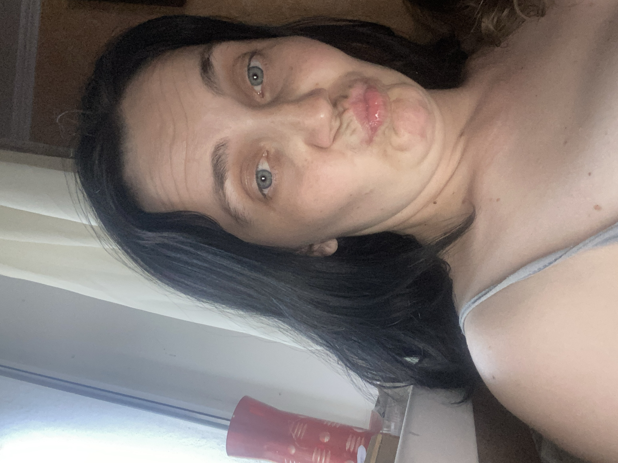 A woman with dark hair and blue eyes puckers her lips for a selfie. She is wearing a light-colored tank top, and her shoulders and chest are visible. Behind her, a window with white curtains is to the left, and a brown wall is to the right. A red vase with a gold pattern sits on a ledge next to the window.