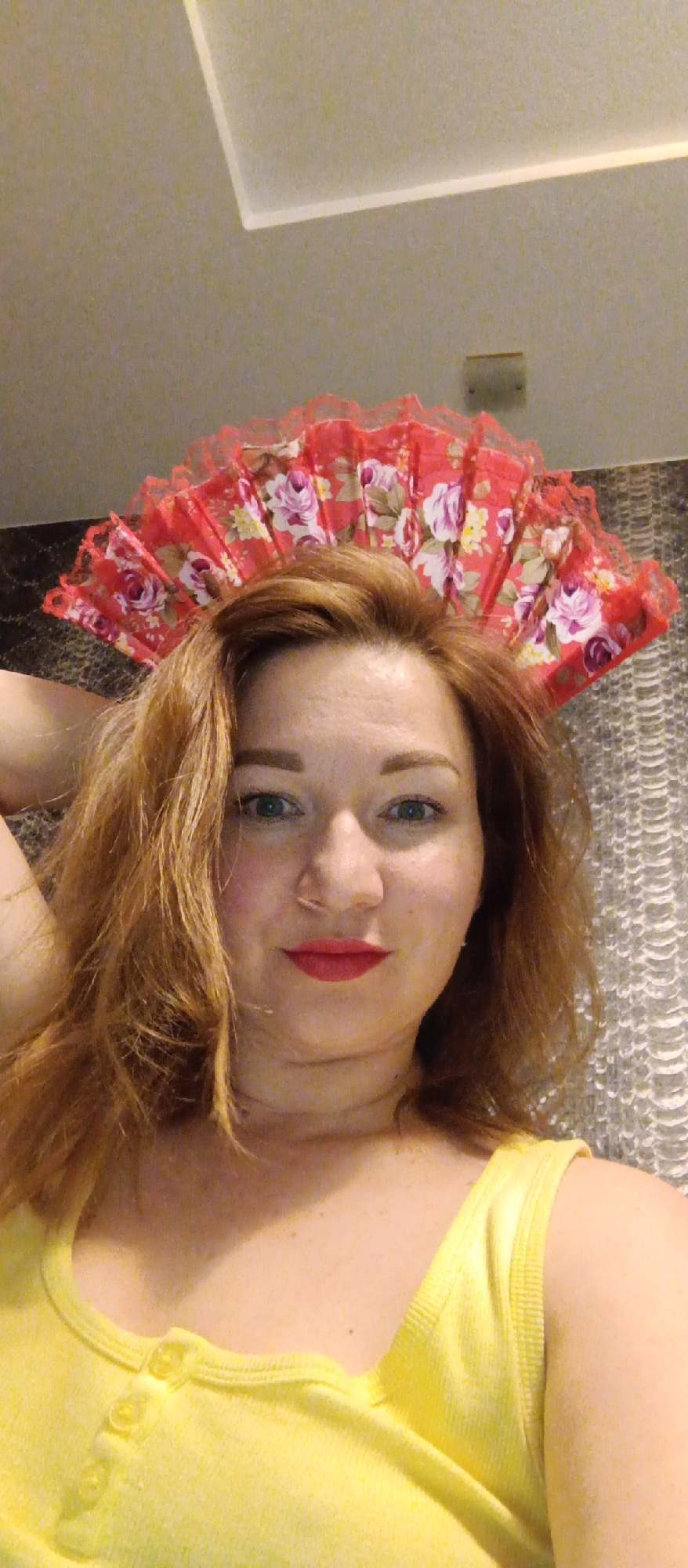 A head-and-shoulders shot of a woman with reddish-brown hair, wearing a yellow tank top. She is smiling slightly with red lipstick. Atop her head is a red fabric fan with a floral pattern of roses and other flowers. The background consists of a gray patterned wall, and the top corner of the frame shows a rectangular lighting fixture on a light-colored ceiling. The location is Unknown, Unknown.