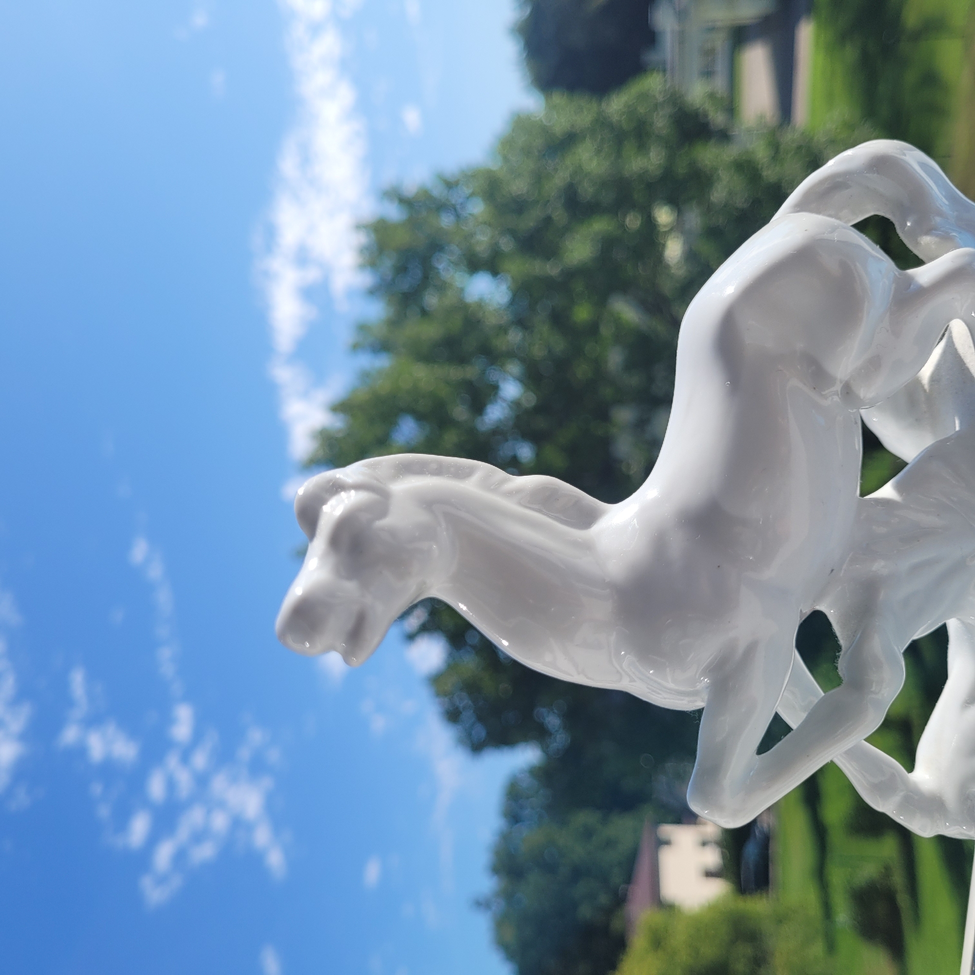 A close-up, eye-level, outdoor, medium shot captures a white porcelain horse figurine in the foreground, its sleek form arcing upwards against a bright blue sky with scattered white clouds. The figurine is glossy, with subtle reflections on its surface. Behind the figurine, a blurred background reveals lush green trees and the distant facade of a building, suggesting a park or garden setting. The specific location in Jūrmala, Latvia, cannot be determined from the visual information provided.