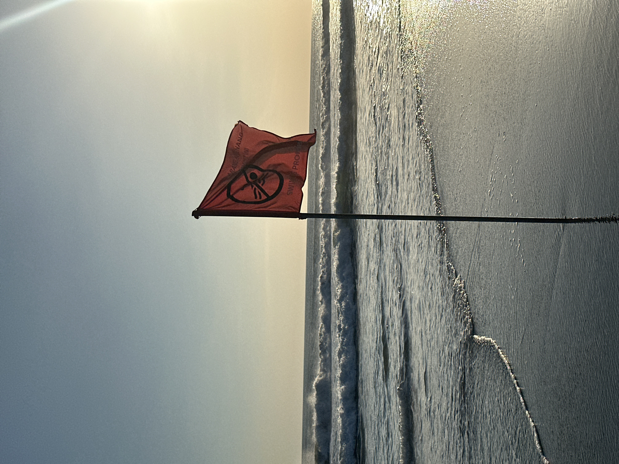 A red flag with a "no swimming" symbol and text is planted in the sand on a beach in North Kuta, Indonesia. The flag is on a dark pole, and the red fabric is billowing in the wind. The ocean is visible behind the flag, with waves crashing on the shore. The sun is low in the sky, casting a warm, golden light over the scene. The sand is wet and reflects the light. The sky is a clear, pale blue.