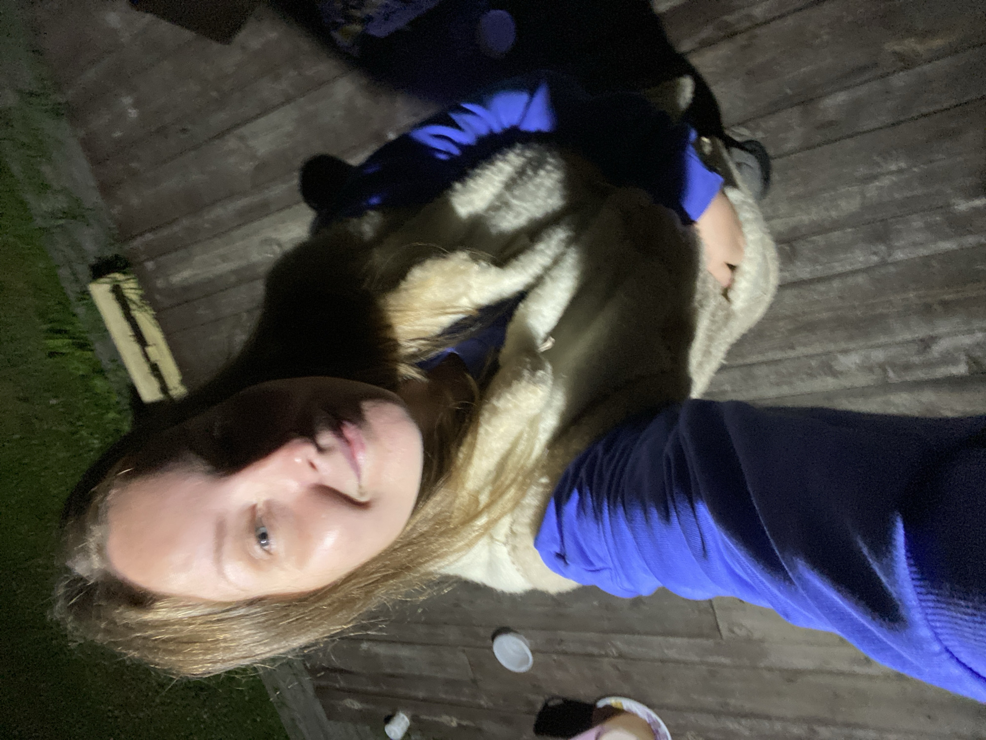 In an outdoor setting at night, on a wooden deck, a person is visible. The individual has light brown hair and is wearing a light-colored fuzzy vest over a blue long-sleeved shirt. The person is holding an object, partially obscured by their vest. The background shows a dark grassy area and more wooden decking. No specific location within the city of Goryaevaya, Russia, can be determined from the provided image.