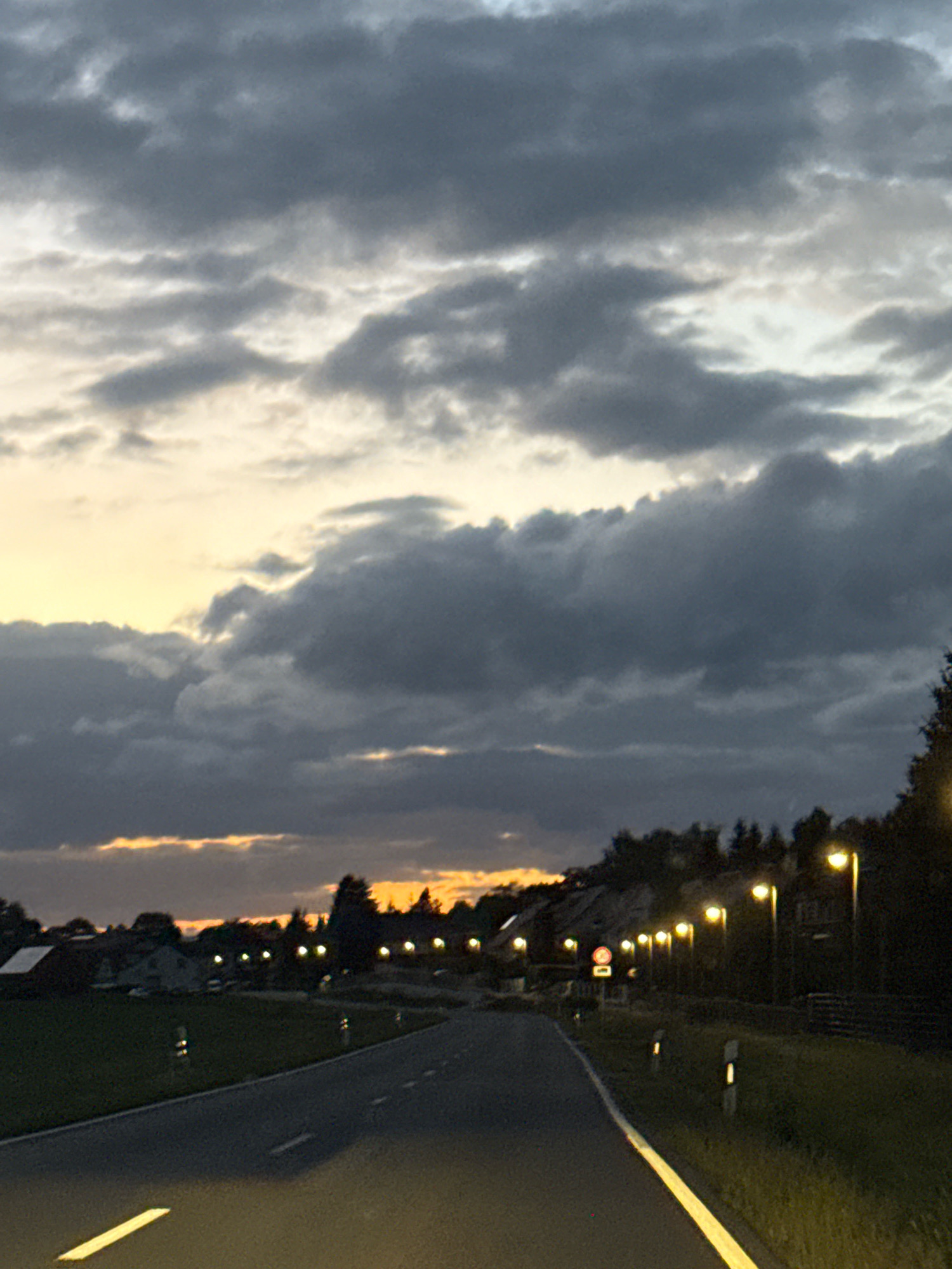 The image captures a road in Werther, Germany, during dusk. The sky is filled with dramatic clouds, ranging from dark gray to lighter tones, with a hint of yellow and orange near the horizon. A paved road, with a yellow lane marker, stretches into the distance. On either side of the road, there are grassy areas with scattered streetlights and some vegetation. In the background, the silhouette of buildings and trees is visible. A few road markers with reflective elements are placed along the roadside. The scene appears quiet and serene, with the artificial lights beginning to illuminate the darkening landscape.