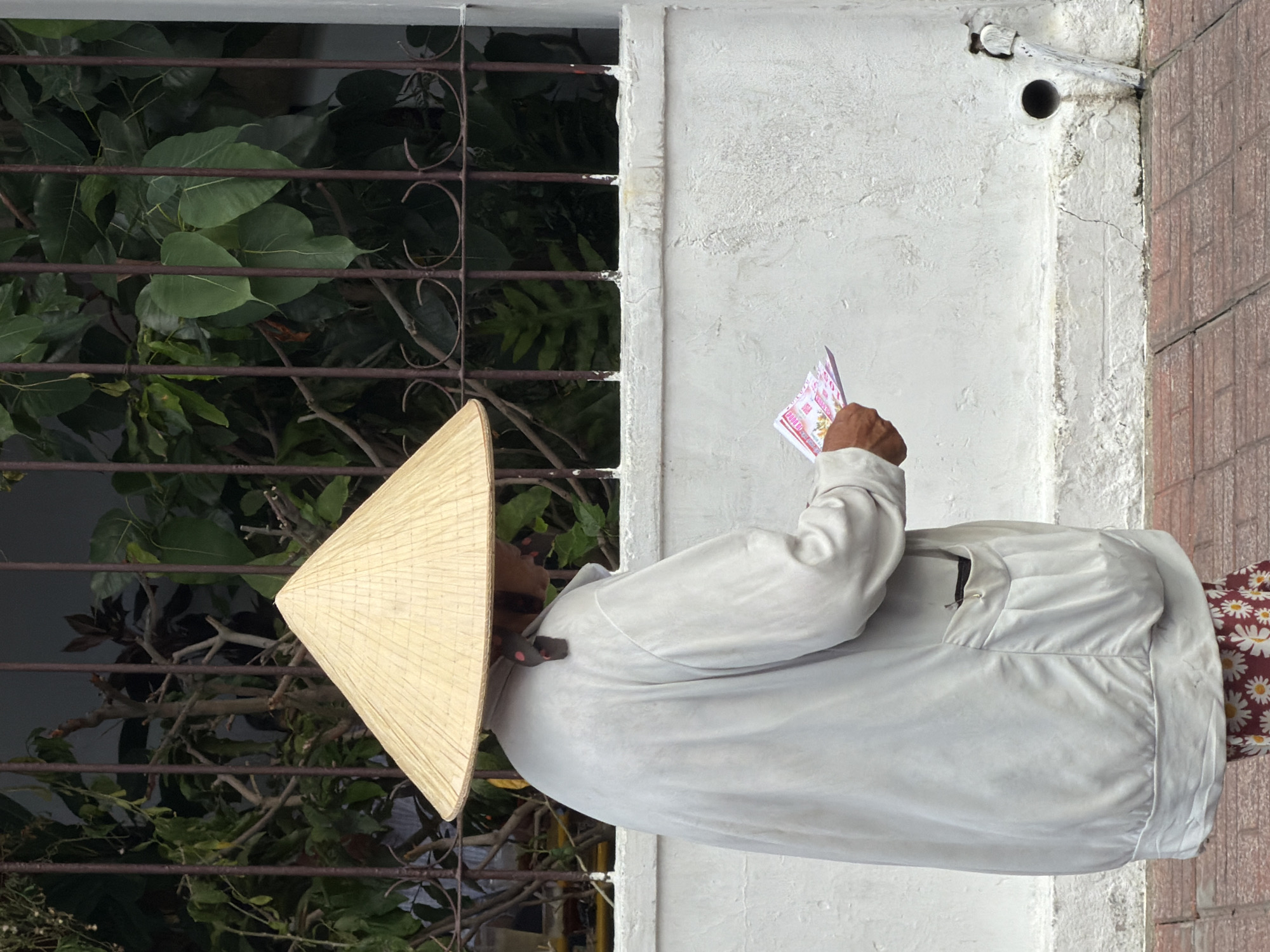 A person wearing a conical hat and a light-colored, loose-fitting jacket is holding a small stack of pink and white folded papers in their right hand. They are standing outdoors in front of a white wall with a decorative metal grate and foliage behind it. The ground is paved with reddish-brown bricks. A drainpipe is visible on the right side of the wall near the ground. The person's lower body is obscured by the jacket, but a portion of a patterned skirt with white flowers on a red background is visible at the bottom of the image. The scene appears to be on a street in Nha Trang, Vietnam.