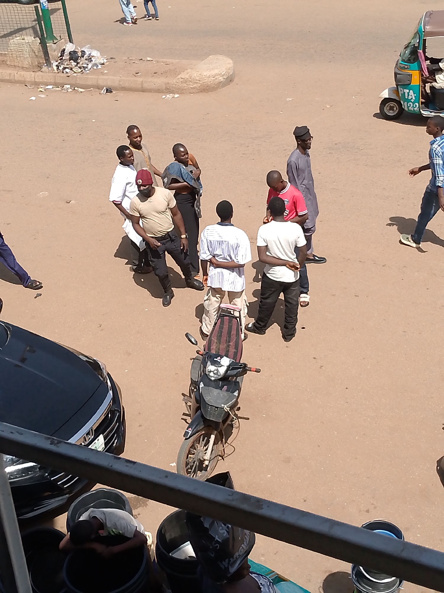 The image depicts a street scene in Bukuru, Nigeria. A group of men are gathered in the middle of the street. Some are wearing traditional clothing, while others wear more casual attire. One man wears a burgundy fez-like cap. A motorcycle is parked nearby. A black car is partially visible on the left side of the image. Trash is scattered on the side of the road. A man appears to be sleeping in a large bucket in the lower portion of the photo. A three-wheeled auto-rickshaw (tuk-tuk) is visible in the upper right.