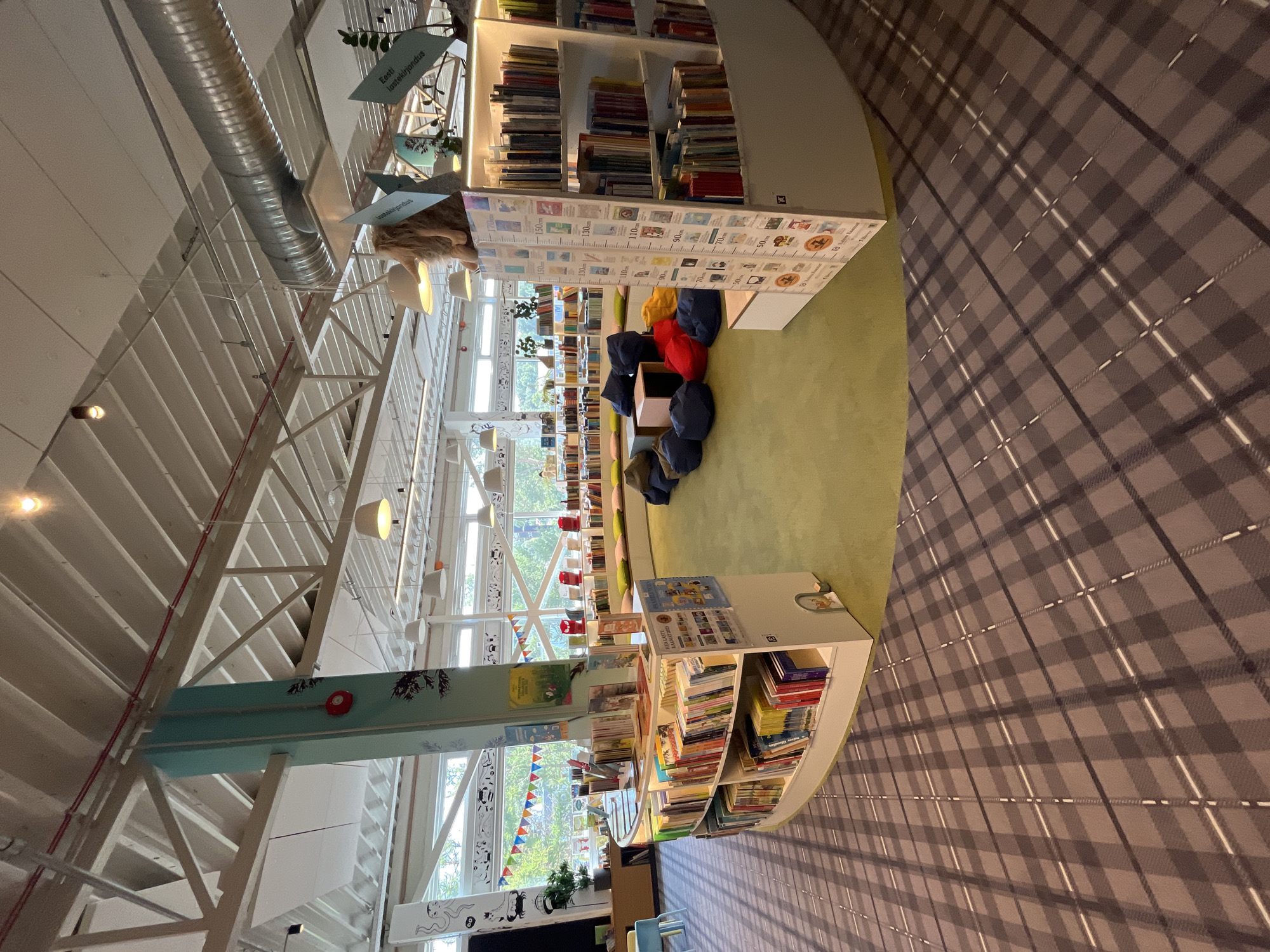 The image shows a well-lit, modern library interior in Haabneeme alevik, Estonia. The library features a central green carpeted area with several large, dark blue beanbag chairs and some red and yellow cushions, creating a comfortable seating nook. Surrounding this area are curved bookshelves filled with a variety of books, some of which are displayed at an angle. A tall, light blue pillar with decorative drawings is a prominent structural element in the center of the space. Large windows offer views of greenery outside, suggesting a pleasant outdoor environment. The ceiling has exposed beams and industrial-style lighting fixtures. The floor in the foreground is covered with a grey and white plaid carpet. On the right side, a tall shelf unit displays more books and a height chart. A small stuffed animal is visible on top of a bookshelf in the background. There are no people visible in the image.