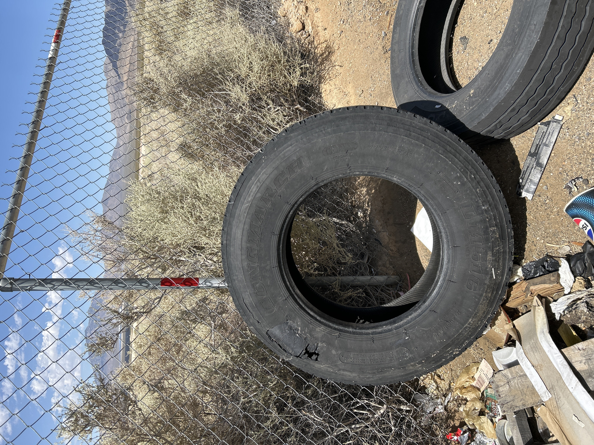 In Primm, Nevada, a photograph captures a scene of abandoned tires and debris near a chain-link fence. The fence, with a metal post adorned by a red and white reflective marker, encloses a dry, arid landscape with sparse, light-brown brush and distant mountains under a clear blue sky with scattered clouds. In the foreground, two large, worn tires lie on the sandy ground. One tire, centrally located and damaged with a large hole in its sidewall, displays the partial text "LONGMAR". Beside it, another tire rests. A pile of discarded items, including wood scraps, plastic bags, and other miscellaneous trash, is visible at the base of the fence and near the tires. The overall impression is one of neglect and disuse in a desert environment.