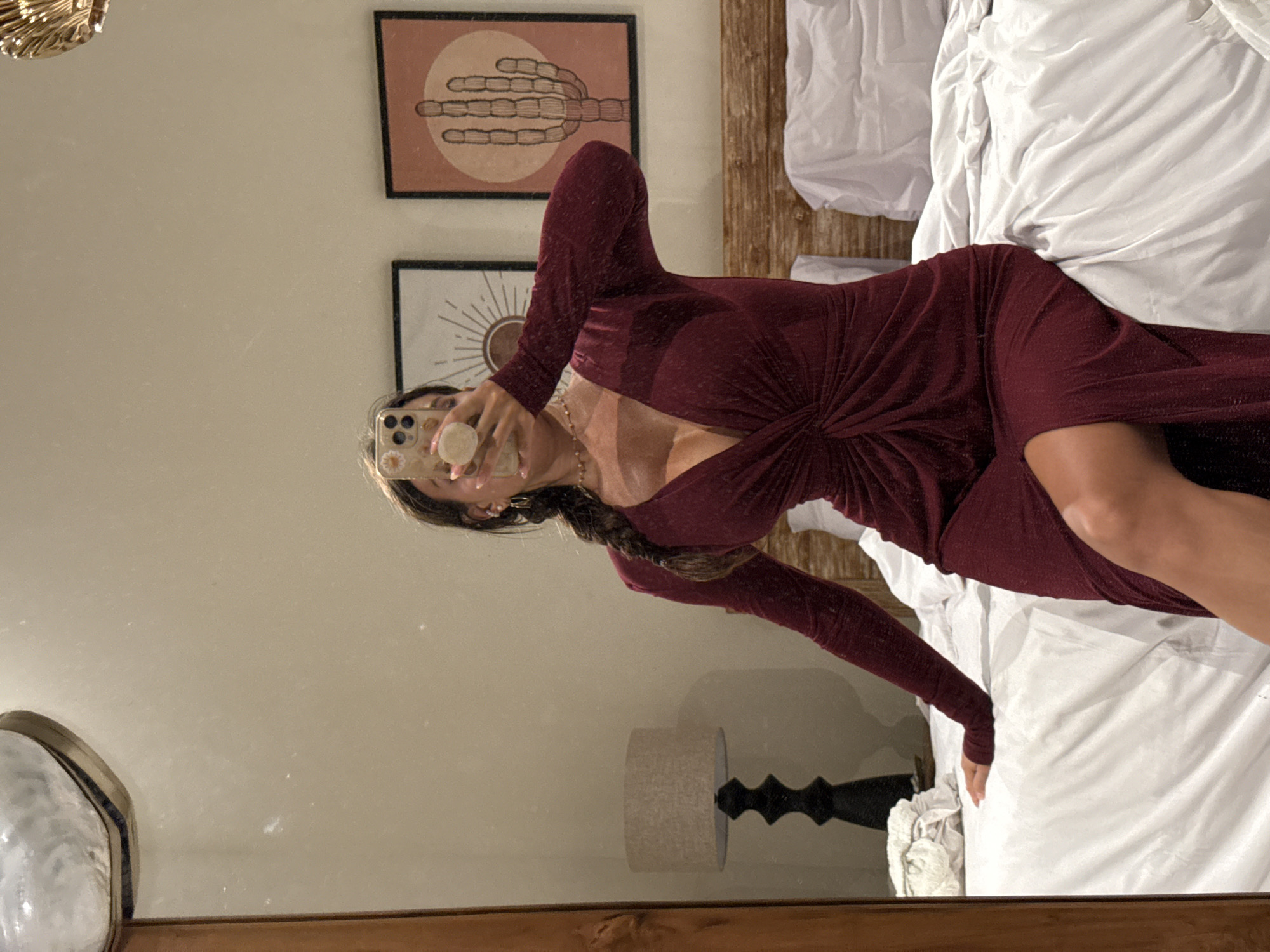 A vertical mirror selfie shows a person reclining on a bed in what appears to be a bedroom in North Kuta, Indonesia. The image is rotated 90 degrees clockwise, giving the impression the person is lying horizontally.
The main subject is wearing a long-sleeved, deep burgundy or maroon maxi dress with a subtle textured pattern. The dress features a plunging V-neckline and a twisted fabric detail at the waist. She has long, dark hair styled in a braid and is wearing a necklace and earrings. Her face is partially obscured by the phone she holds up to take the picture. The phone case is light-colored with small floral designs, and it has a white pop-socket attached. Her fingernails are painted a light shade.
Reflected in the mirror, behind the subject, is a light-colored wall adorned with two framed art prints. The upper print depicts a stylized cactus or similar plant form against an orange circular background. Below it, the second print features a sunburst or radial pattern. To the right of the reflection, a bed with white, ruffled bedding and a rustic wooden plank headboard is visible. A bedside table holding a lamp with a light-colored cylindrical shade and a dark, sculptural base is also reflected in the lower part of the mirror. The mirror surface shows some dust or smudges. In the top left corner of the original image, outside the mirror's reflection, part of a golden, ornate object is visible. The scene suggests a relaxed, indoor setting. No visible text is present.