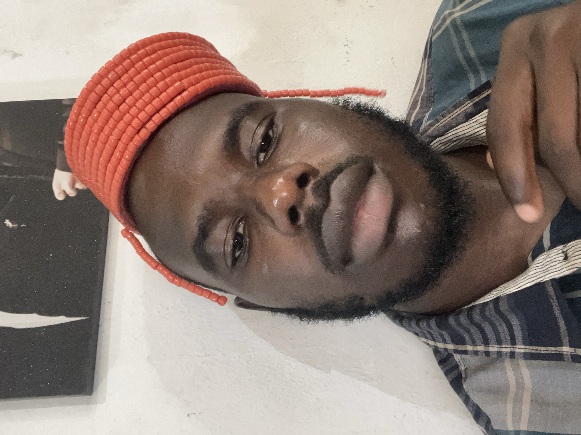 A medium shot captures a Black male from the chest up, looking directly at the camera. He wears a red beaded hat and a blue and gray plaid shirt. The background consists of a white, textured wall and a black and white framed picture hanging on it. The man has a beard and appears to be indoors. The image quality is moderate, with some graininess and lighting that creates shadows. There are no identifiable landmarks that would indicate a specific location within Jos, Nigeria.
