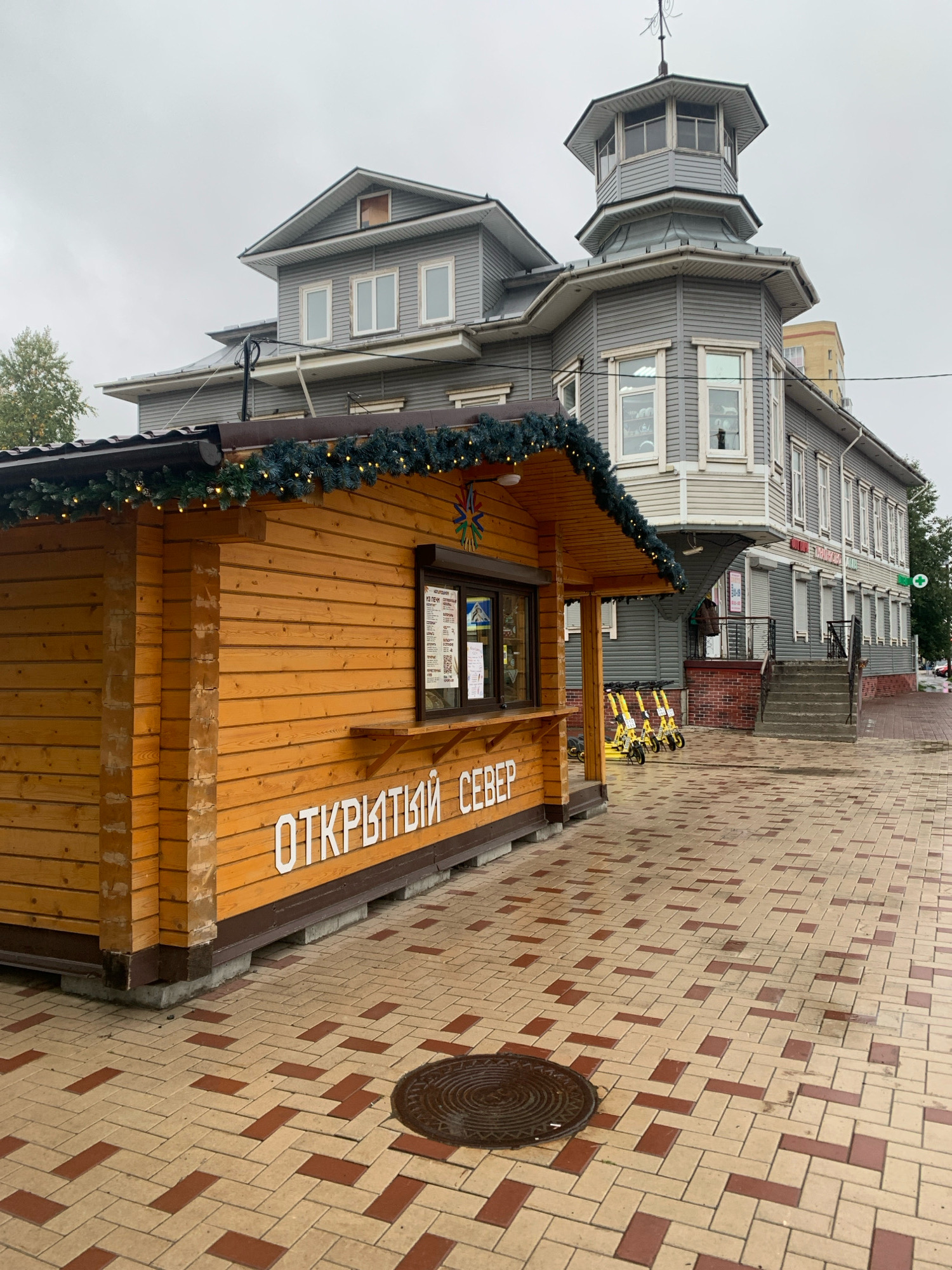 A wooden kiosk with the words "ОТКРЫТЫЙ СЕВЕР" (Open North) painted in white lettering is positioned in the foreground, to the left. The kiosk is decorated with a garland of evergreen branches and small lights. Behind the kiosk, a large, multi-story gray building with a distinctive octagonal tower on the right corner stands. The building features numerous windows and is clad in horizontal siding. Yellow electric scooters are parked in a row to the right of the kiosk. The ground is paved with light and dark red rectangular bricks laid in a herringbone pattern, and a circular manhole cover is visible on the pavement. The sky is overcast and gray. The scene appears to be a street or public area in Arkhangelsk, Russia.