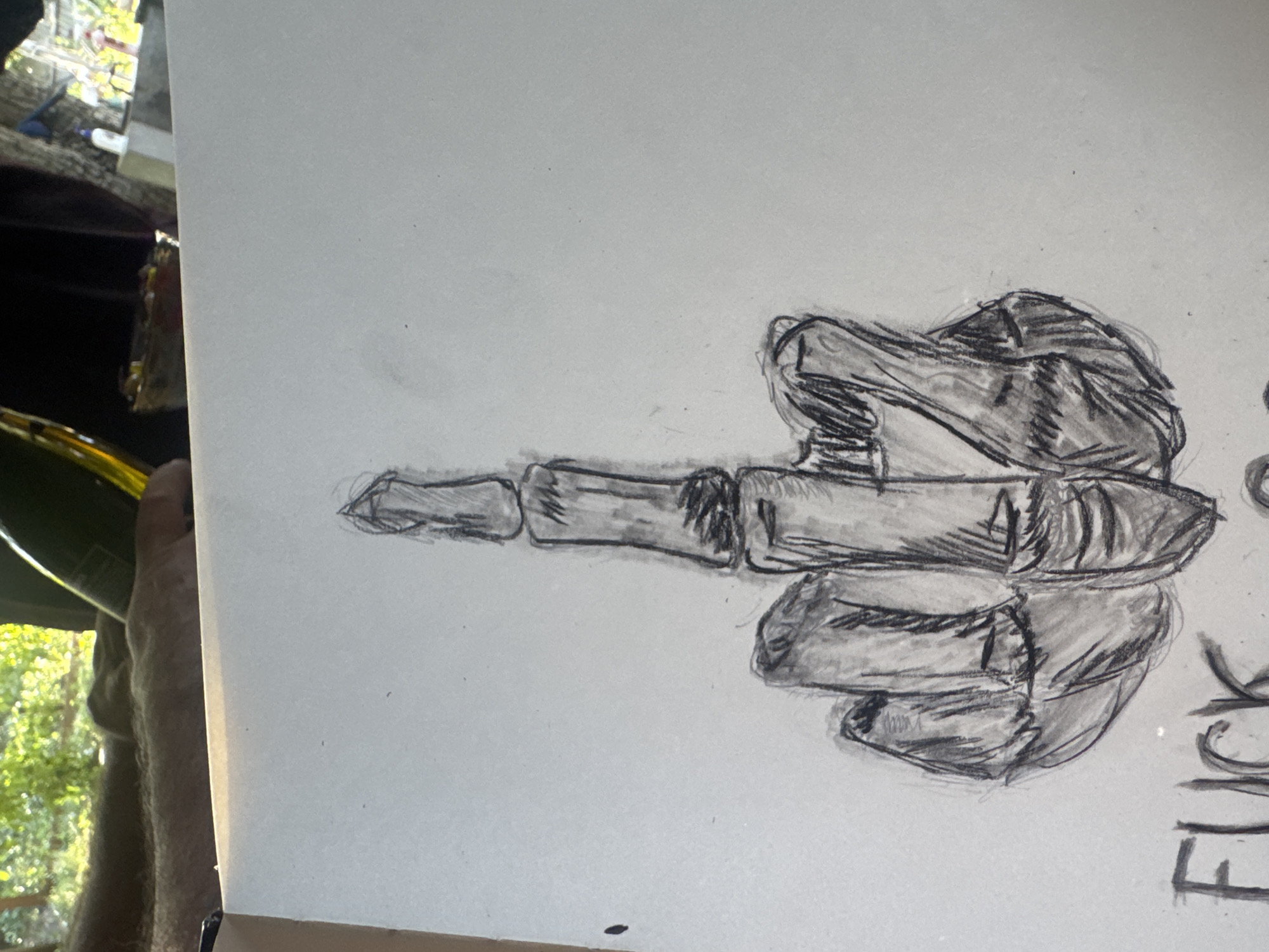 A charcoal sketch on a white paper is shown. The sketch depicts a skeletal middle finger raised, with other fingers curled into a fist. The word "FUCK" is written in capital letters in the lower left corner of the paper. The sketch is partially obscured by a blurred background that shows a person's arm and a lush green environment, possibly a garden or trees. The scene is indoors, with natural light coming from an unseen window. The location is not specified in the image or its metadata.
