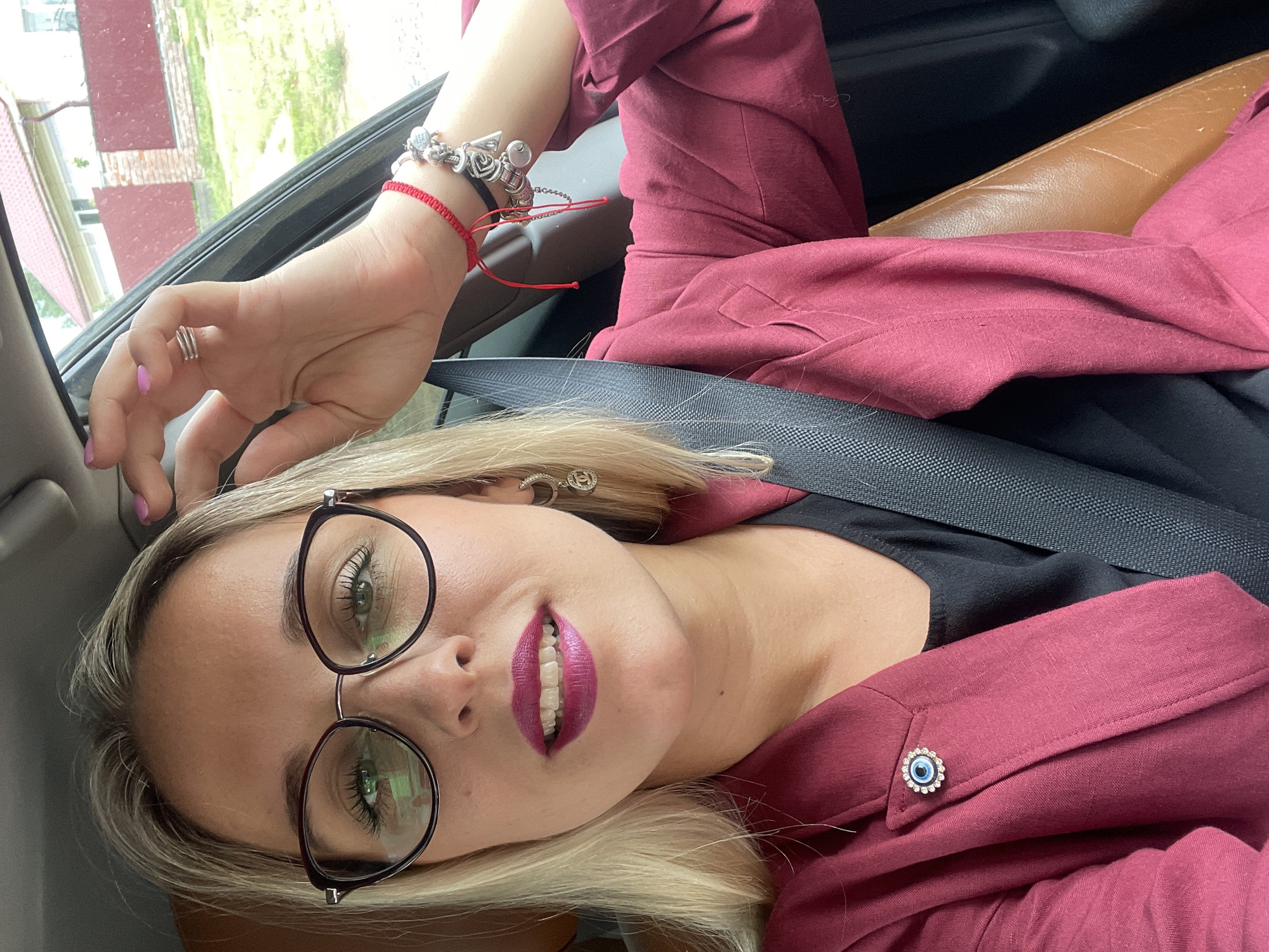 The media file is a selfie taken from the perspective of someone sitting in the passenger seat of a car. The main subject is a young woman with blonde hair, wearing glasses, a maroon blazer over a black top, and makeup. She is wearing several pieces of jewelry, including a ring on her left hand and a bracelet with charms on her wrist. A red string bracelet is also visible on her wrist. A small, circular brooch with a blue eye design is attached to her blazer. The car's interior is visible, including a brown leather seat and a black seatbelt. Through the car window, a blurred background of buildings and greenery can be seen, suggesting an outdoor setting. The woman is smiling and looking at the camera, conveying a sense of happiness and confidence. The lighting suggests it is daytime. No visible text is present in the image. The location context is Chaikovsky, Russia.