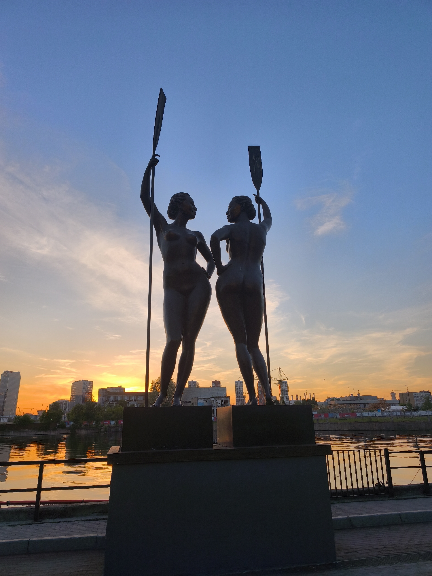 This image captures a wide shot of two bronze statues depicting nude female figures, known as "The Oarswomen" or "The Rowers," prominently displayed on the Frunzenskaya Embankment alongside the Moskva River in Moscow, Russia. The scene is set during a sunset, with the sun positioned low on the horizon behind the statues.
The statue on the left faces towards the right, its right arm extended upwards, holding a paddle vertically. Its silhouette reveals the torso and upper thighs. The statue on the right faces away from the viewer, also positioned towards the right, with its left arm raised, holding a paddle. This figure is seen in full, from behind, emphasizing its muscular form. Both statues stand on dark, rectangular pedestals.
The sky exhibits a dramatic gradient, transitioning from an intense orange and yellow at the horizon, where the sun's glow is brightest, to a clear, deep blue in the upper sky, interspersed with wispy white clouds. The Moskva River in the middle ground reflects the vibrant sunset colors, creating shimmering streaks of orange and gold on its surface.
Across the river, a modern cityscape is silhouetted against the bright horizon, featuring multiple high-rise buildings and several construction cranes, indicating ongoing urban development. In the immediate foreground, a dark metal railing runs parallel to the riverbank, with a segment of a paved walkway visible between the railing and the statue pedestals. The statues themselves are heavily backlit by the setting sun, rendering them as dark, almost monochromatic silhouettes against the vivid sky.