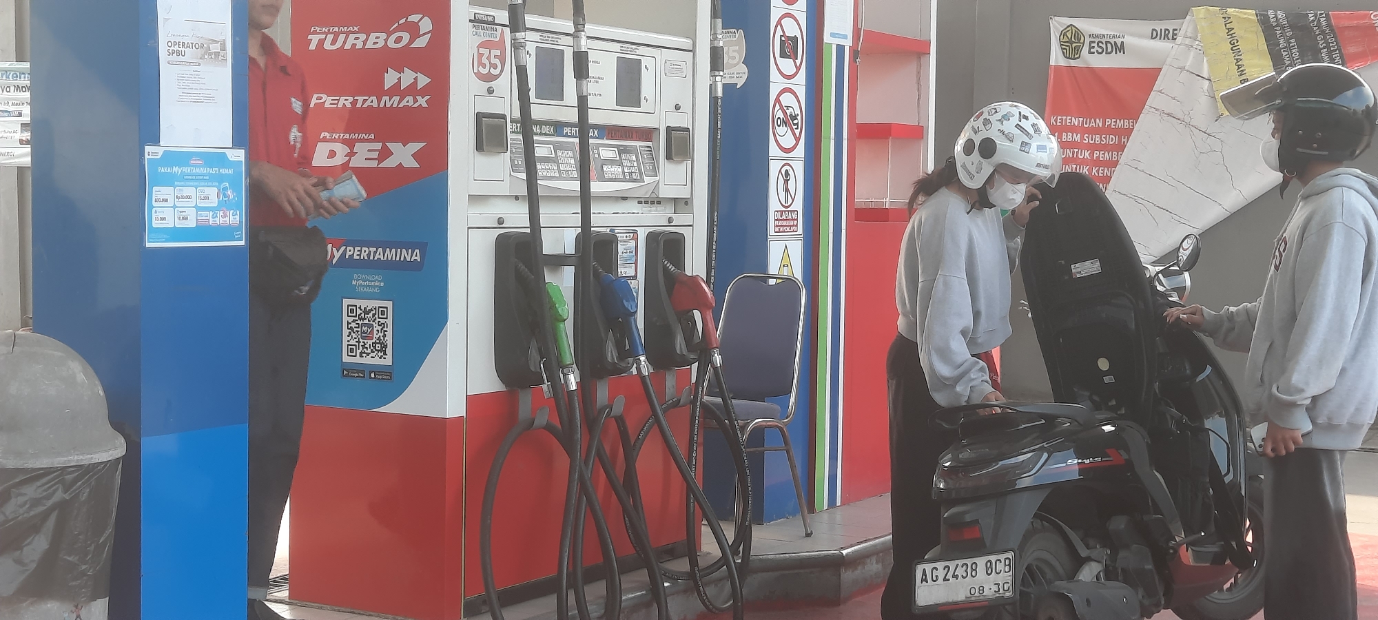 In Lodoyo, Indonesia, a Pertamina gas station scene unfolds. A gas pump unit displays "PERTAMAX TURBO," "PERTAMAX," and "DEX." A male attendant, wearing a red uniform and holding cash, stands to the left of the pumps. Two individuals, one with a white helmet covered in stickers and the other with a black helmet, are near a black scooter with license plate "AG 2438 OCB". They appear to be interacting around the scooter. The environment includes a trash bin, a blue chair, and posters with text including "OPERATOR SPBU" and "KEMENTERIAN ESDM".