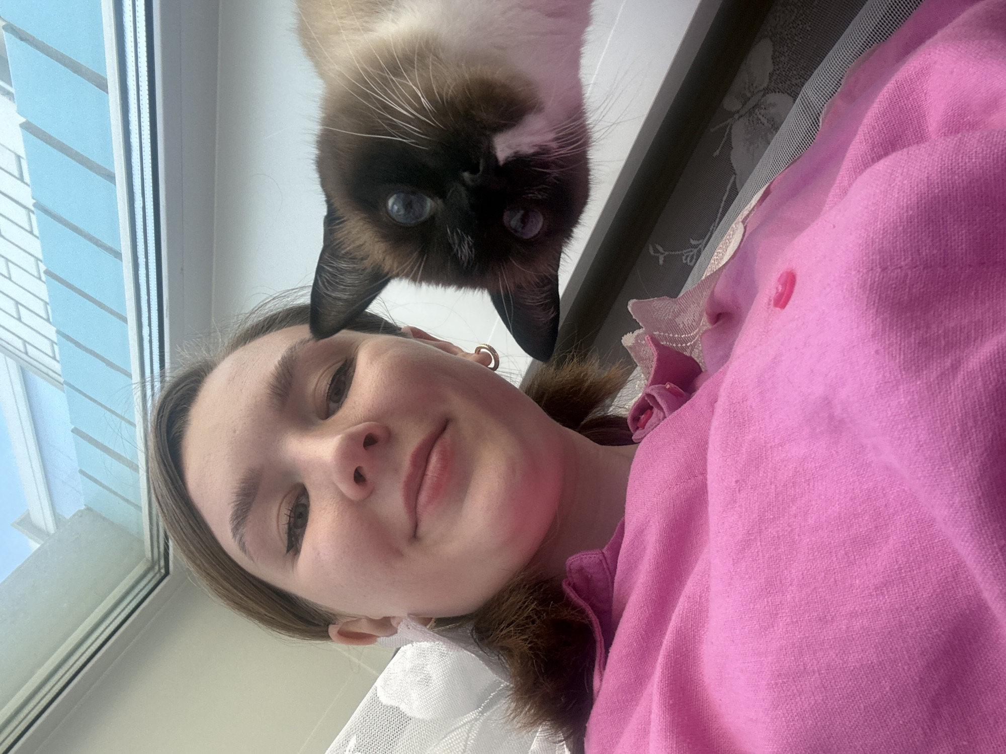 A selfie-style photograph, captured from a low angle, features a young woman and a Siamese cat. The woman, with fair skin and brown hair, is wearing a pink top. Her face is centered in the lower half of the frame. Above her, the Siamese cat is positioned with its face directly above hers, peering down. The cat's dark brown and white fur is visible. The background includes a window on the left side of the frame, revealing a glimpse of what appears to be outdoor lighting. A patterned surface is partially visible in the upper right corner, possibly a wall or a part of the interior. The location is unknown and there is no information available to confirm the location in the city of Izhevsk, Russia.