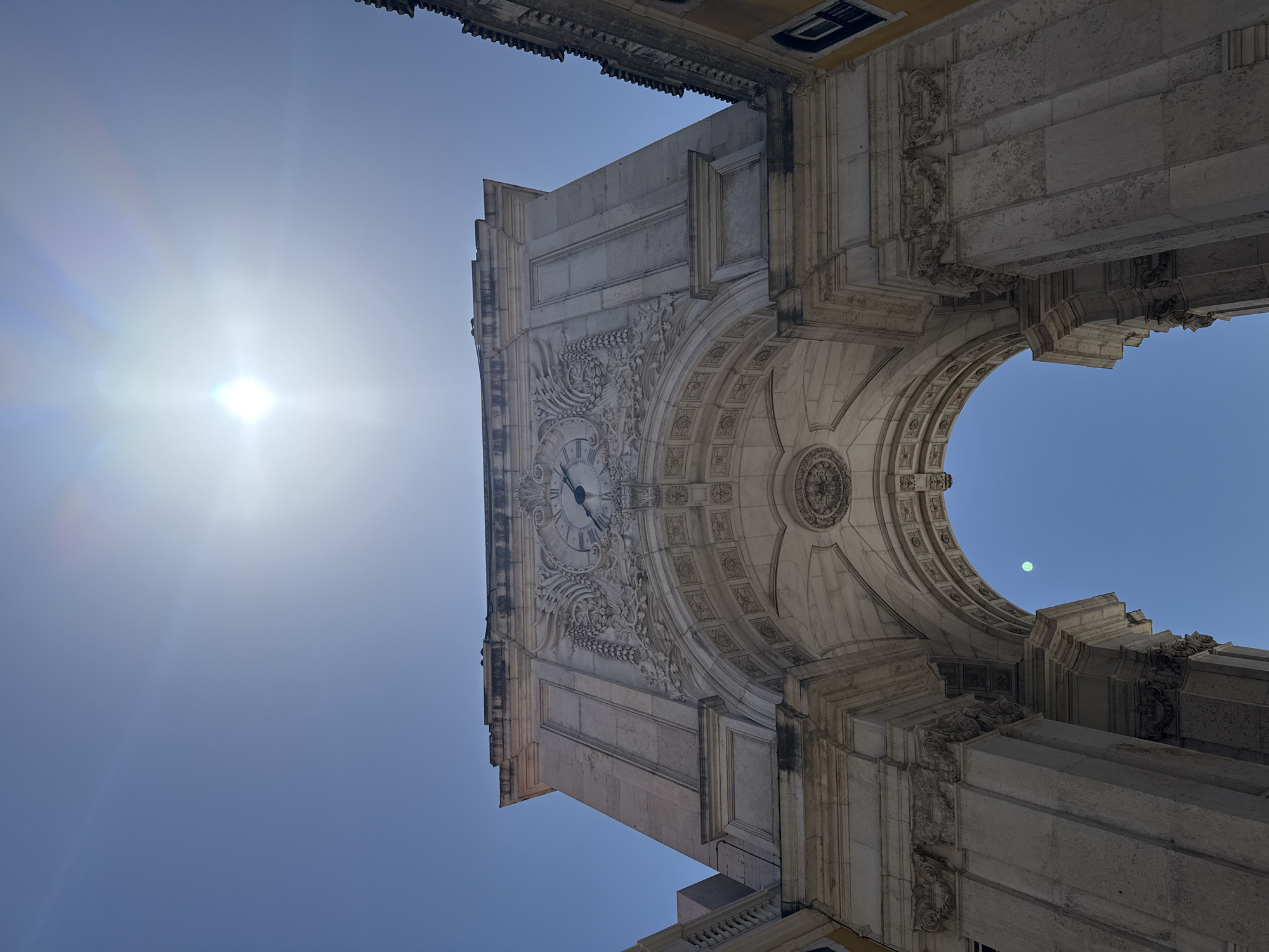 The image shows a low-angle, exterior view of the Rua Augusta Arch in Lisbon, Portugal, against a clear blue sky with the sun prominently featured and creating lens flare. The ornate stone archway is made of light-colored stone and features intricate carvings, including a large clock face above the arch. The arch is flanked by two buildings, one on the right with a yellow facade and a dark-tiled roof, and another on the left which is partially visible. The architecture suggests a grand, historic urban setting. There are no people visible in the image.