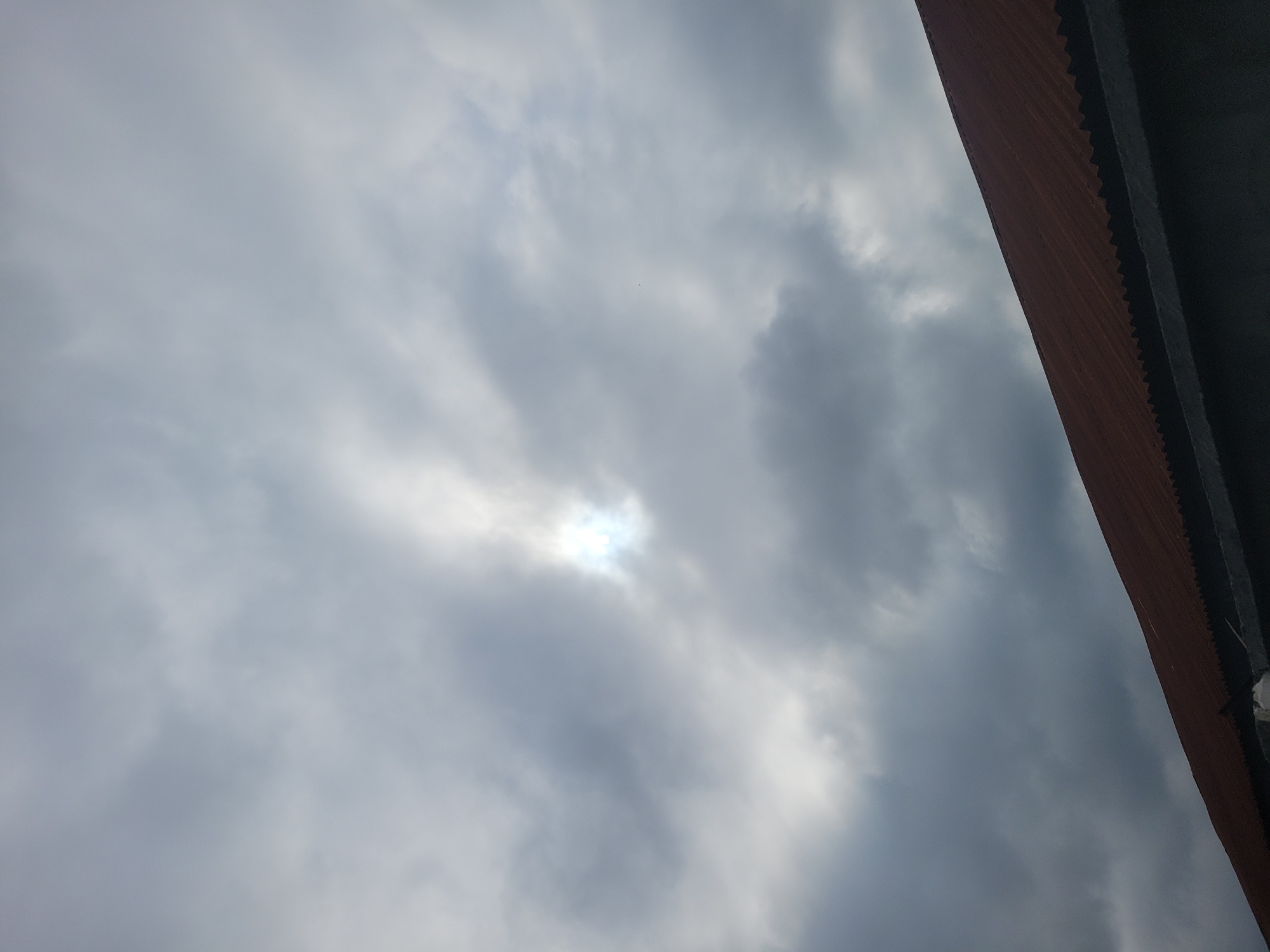The image shows a cloudy sky with the sun partially visible through a break in the clouds. The lower portion of the image is occupied by the corrugated metal roof of a building, which is reddish-brown in color. A dark grey concrete structure runs along the bottom edge of the image, below the roof. The scene appears to be outdoors, possibly in an urban or suburban environment given the presence of a building. The overall mood of the image is somber due to the overcast sky.