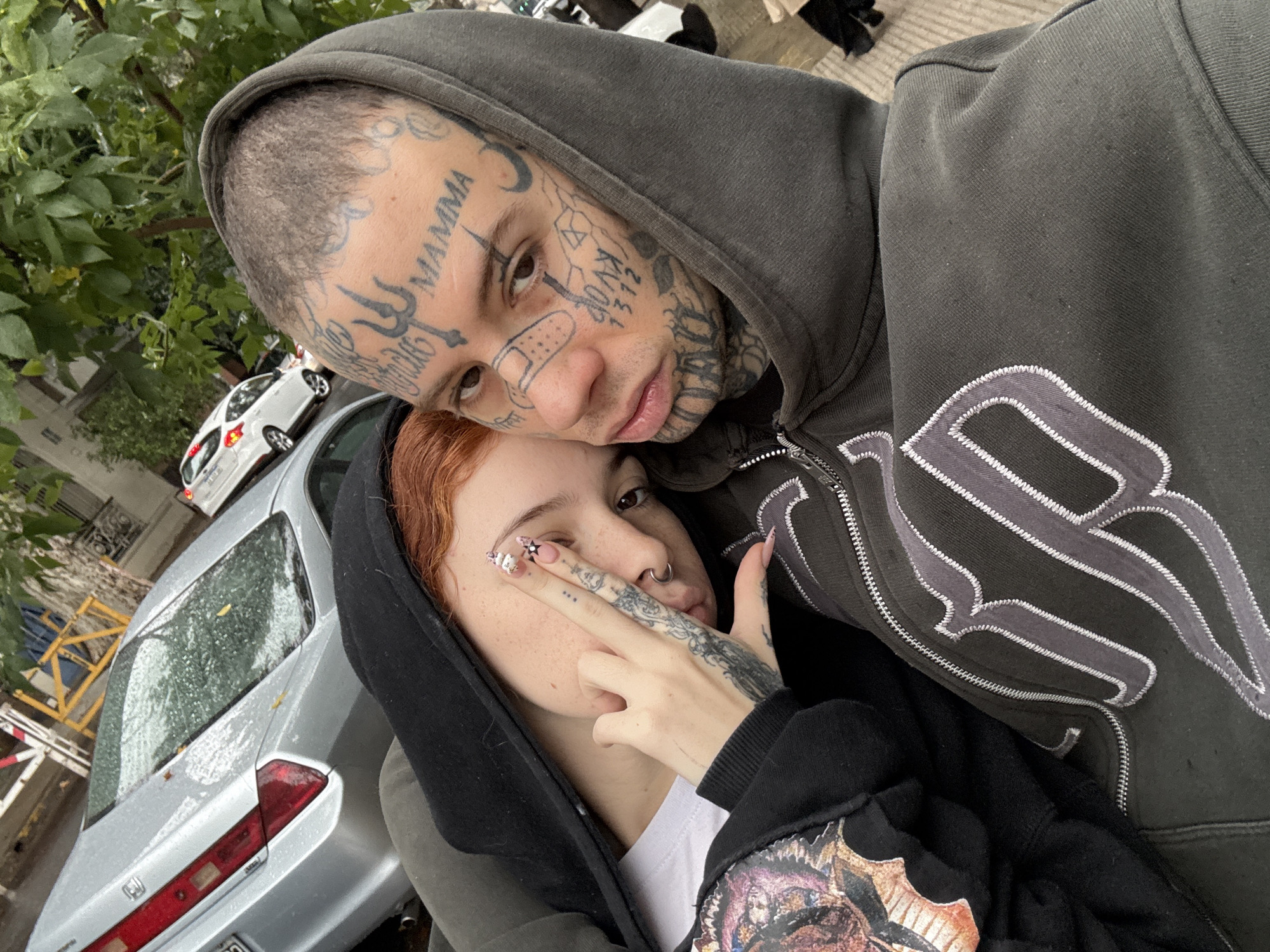 The image shows two people posing for a selfie outside, likely in an urban environment. A person with multiple face tattoos and a shaved head is wearing a grey hooded jacket and leans towards a person with reddish-brown hair, who is wearing a black hooded jacket and has tattoos on their hand. The person with reddish-brown hair holds up their fingers in a gesture. Behind them is a silver car, and the background includes trees and buildings. The location is most likely in an urban environment. Buenos Aires, Argentina is NOT a discernable location.