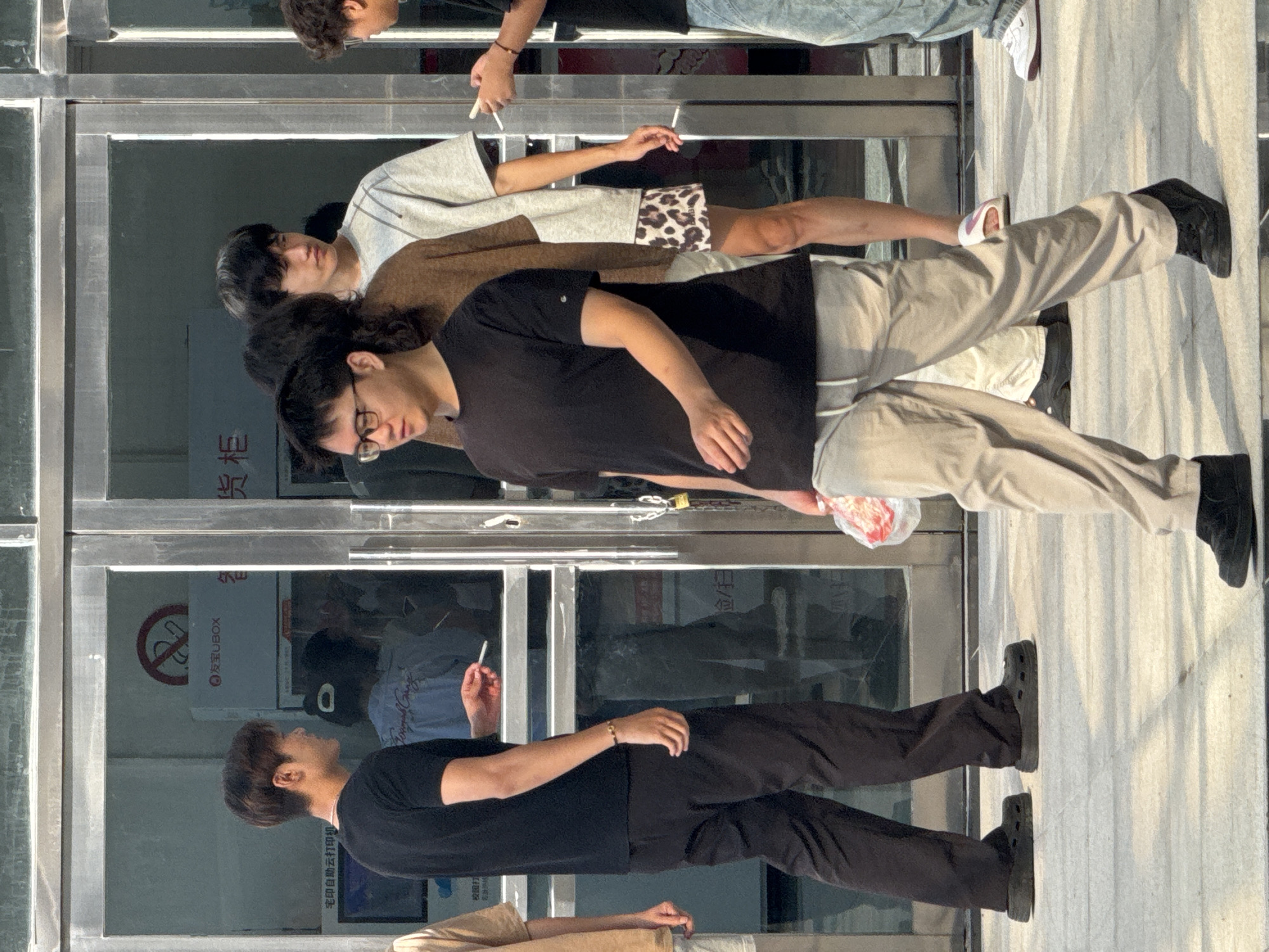 The image depicts a street scene in Guta, China, with several young men standing and walking near a building with glass doors. In the foreground, a man wearing glasses and a black t-shirt walks toward the camera, carrying a plastic bag in his right hand. He is wearing light-colored pants and black shoes. To his left, another man in a black t-shirt and dark pants stands with a cigarette in his right hand, looking towards the left. Behind them, several other young men are visible. One man wears a light gray t-shirt and patterned shorts, another wears a brown shirt, and a third is partially visible on the far right. The background shows the entrance to a building with reflective glass doors and some signage, including a "no smoking" symbol. The ground is paved with concrete or stone tiles. The lighting suggests it is daytime, with sunlight casting shadows on the ground.