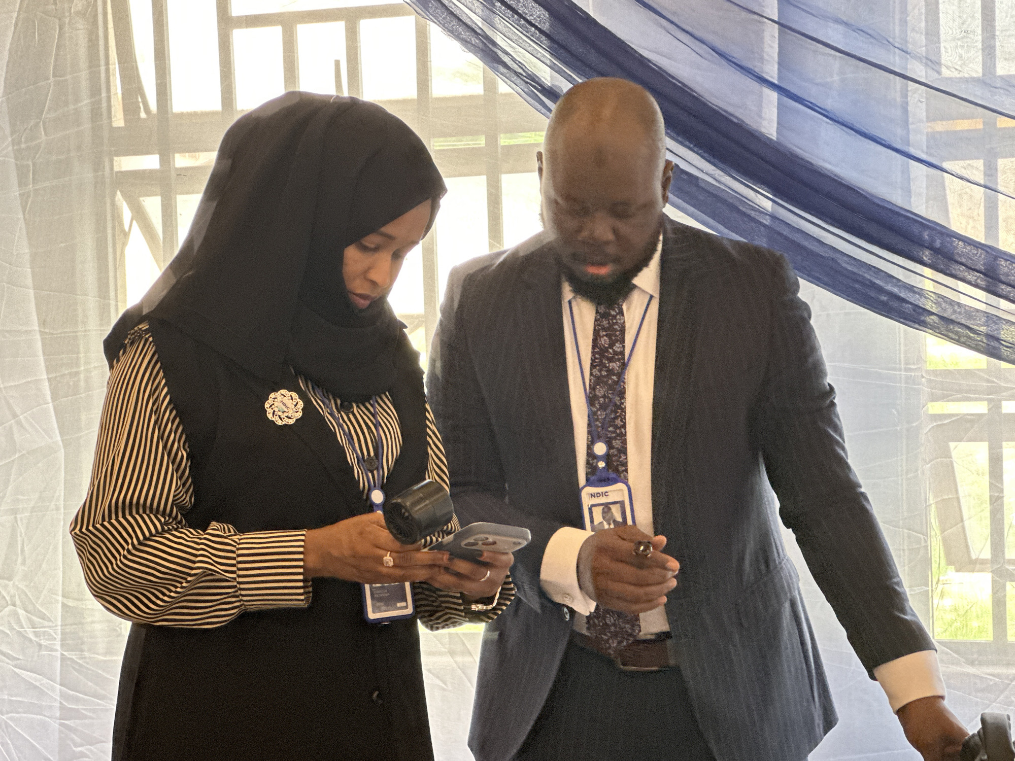 Two individuals, a woman and a man, are captured in a medium shot indoors during the day, likely in Unguwan Dosa, Nigeria. Both are focused downwards, seemingly engaged in an activity involving electronic devices.
The woman, positioned on the left, wears a black hijab that covers her head and neck. She has a striped long-sleeved shirt with black and white vertical stripes, topped by a dark, possibly black, vest or dress. A decorative white brooch is pinned to her attire on her left side. She wears an ID badge around her neck, with part of the blue lanyard visible. In her hands, she holds a light-colored smartphone and a small black cylindrical object, possibly a microphone or scanner. Rings are visible on her fingers.
The man, on the right, is balding and wears a dark pinstriped suit, a white dress shirt, and a patterned tie with shades of blue and purple. He also wears an ID badge around his neck, suspended by a blue lanyard. His badge clearly shows his photo and the text "NDIC." He appears to be holding a pen or a stylus in his right hand. A dark ring is visible on one of his fingers.
The background features large windows with bright light filtering through sheer white curtains on the left and a draped blue, textured sheer curtain on the right. The bright light indicates it is daytime. The scene suggests an indoor event or professional gathering where both individuals are intently reviewing information or using equipment. Their expressions are focused and serious.