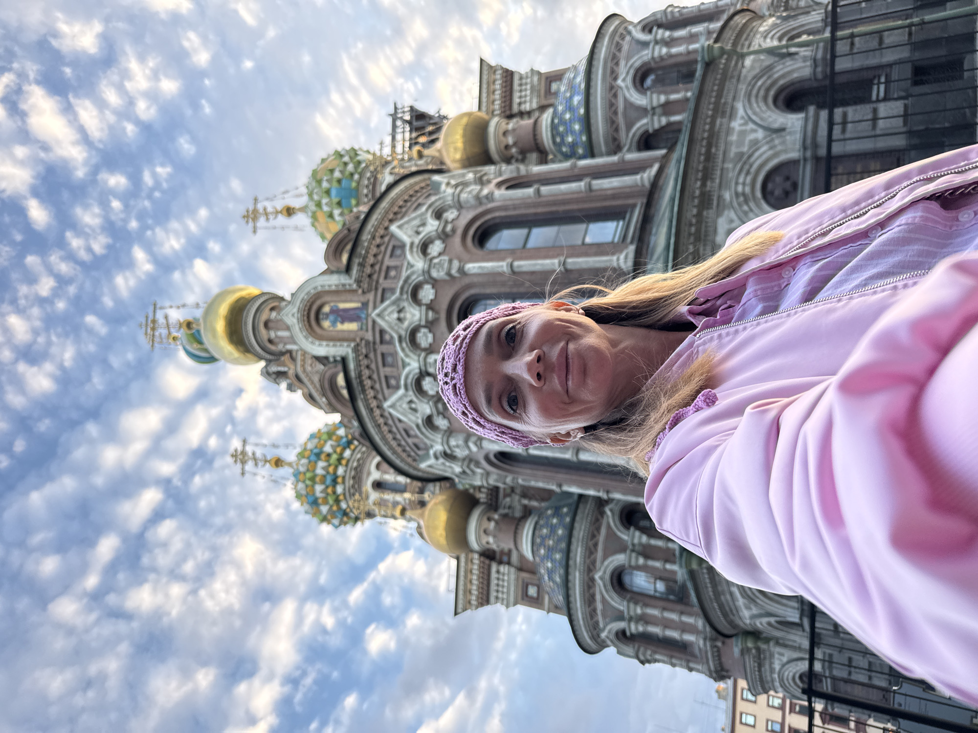 A low-angle, wide-shot selfie of a woman in a pink jacket and a pink knitted headband is taken in front of the Church of the Savior on Spilled Blood in Saint Petersburg, Russia. The woman is smiling at the camera, and her face is illuminated by the soft light of the sky. The church, with its distinctive onion domes and intricate architecture, is visible in the background. The sky is filled with fluffy white clouds, adding a dramatic touch to the scene. The image is captured from a low perspective, making the church appear grand and imposing.