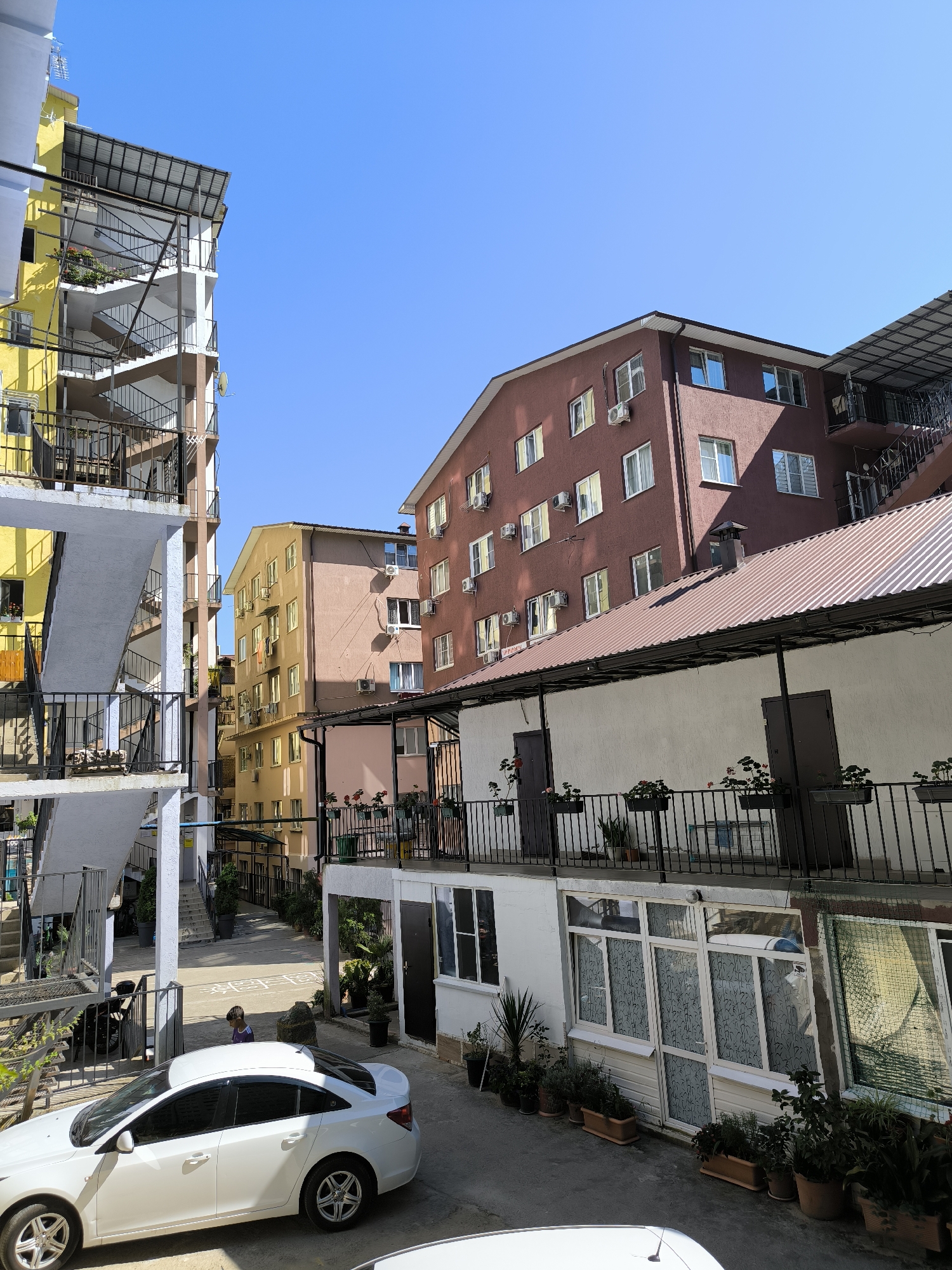 This is a daytime, eye-level, outdoor shot of an apartment complex in Sochi, Russia, with multiple buildings of varying heights and colors. The architecture features external staircases and balconies with black metal railings. The buildings are painted in shades of yellow, peach, and a dark reddish-brown. The sky is clear and blue. In the foreground, a white sedan is parked on an asphalt surface. A child in a purple and blue shirt is visible in the middle ground, near a hopscotch grid drawn on the ground. Several potted plants are arranged on balconies and along the ground floor. The scene depicts a residential area with multiple stories and a clear, sunny day.