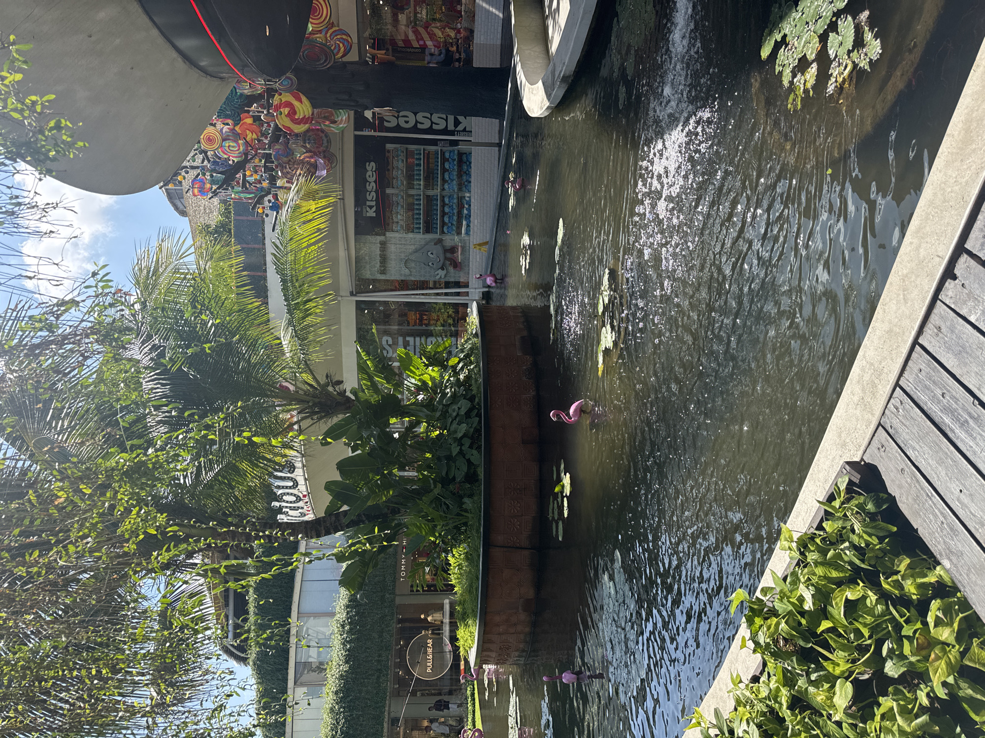 The image shows an outdoor courtyard with a central pond. The pond contains several pink inflatable flamingos. Lily pads are floating on the surface of the water. In the background, there are several buildings with shops. One shop is labeled "Pull&Bear" and another "Tommy Hilfiger." A shop labeled "Kisses" is visible, featuring large, colorful candy-like decorations. Lush green plants and palm trees are present throughout the courtyard. The scene appears to be a shopping center or a public square in Kuta, Indonesia, given the presence of popular retail brands and tropical vegetation. The sun is shining, casting reflections on the water.