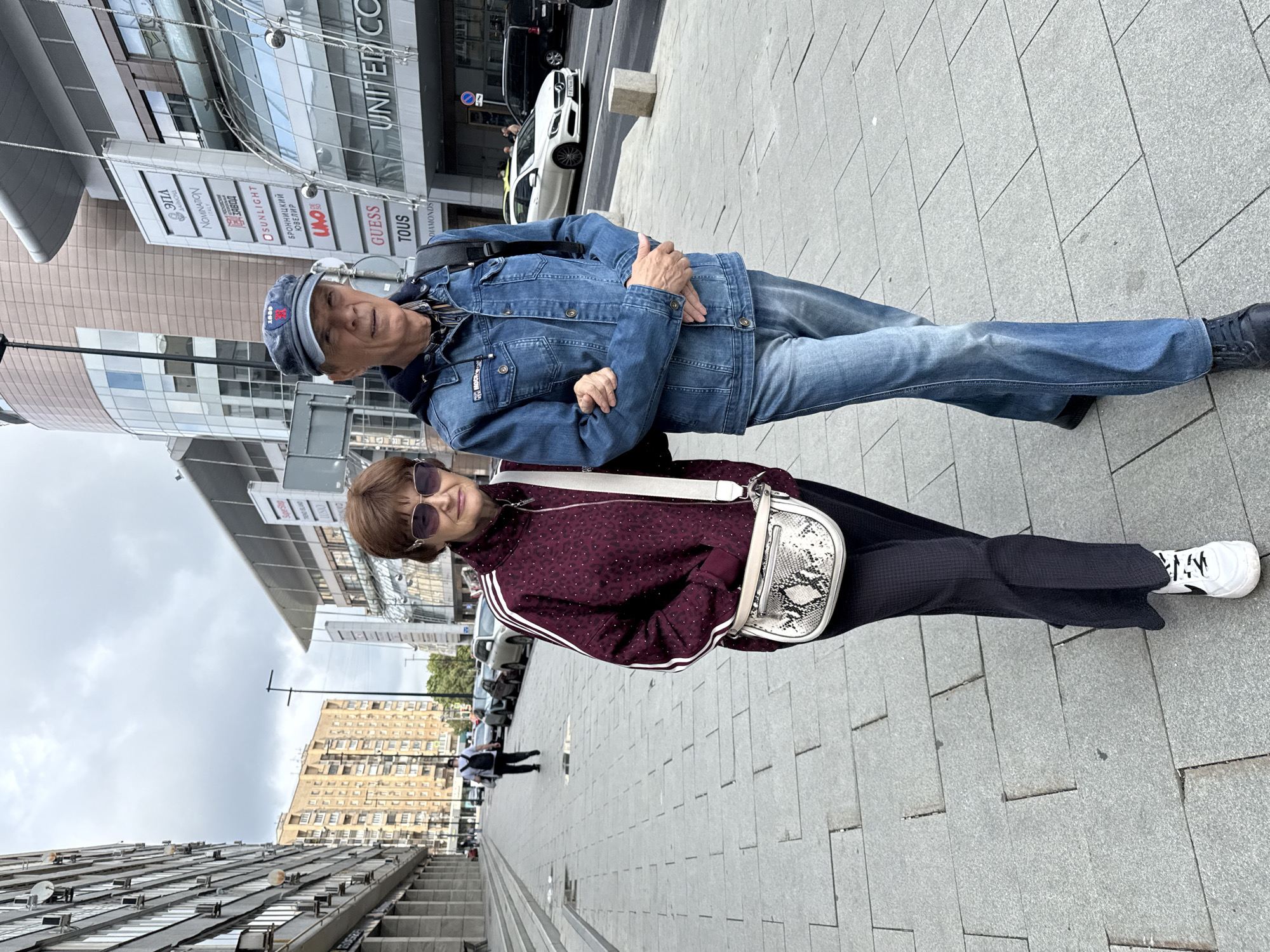 A middle-aged couple walks arm-in-arm on a wide, grey paved sidewalk in front of a modern shopping mall in Moscow, Russia. The man, wearing a blue denim jacket, jeans, and a baseball cap, has his left arm around the woman's waist. The woman, in a patterned maroon jacket and dark trousers, carries a white cross-body bag with a snakeskin pattern. Behind them, several cars are parked along the street. The mall features large glass windows and vertical signage with various brand names, including "UNITED CO". The architecture is contemporary, with clean lines and an emphasis on glass and metal. The sky is overcast. The scene appears to be captured during daytime.