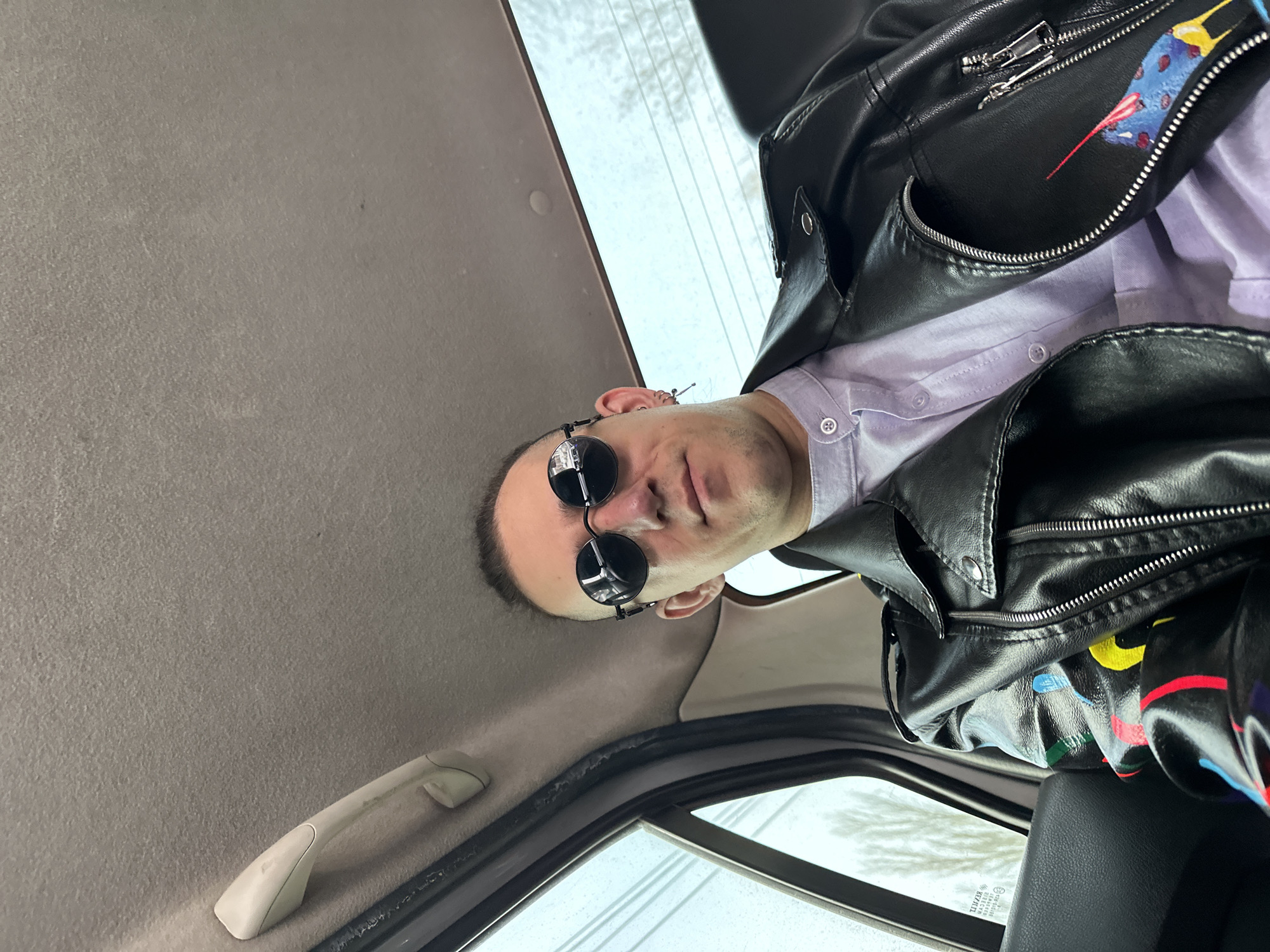 The photo is taken from a low angle inside a car. A person wearing a light purple button-down shirt and a black leather jacket with colorful artwork is looking directly at the camera. The person is also wearing round sunglasses. The car's interior is visible, including the gray roof lining, a grab handle, and a window. Outside the window, it appears to be a cloudy or overcast day. The location is unknown, and nothing in the image allows to determine the city of Saratov, Russia, as the location.