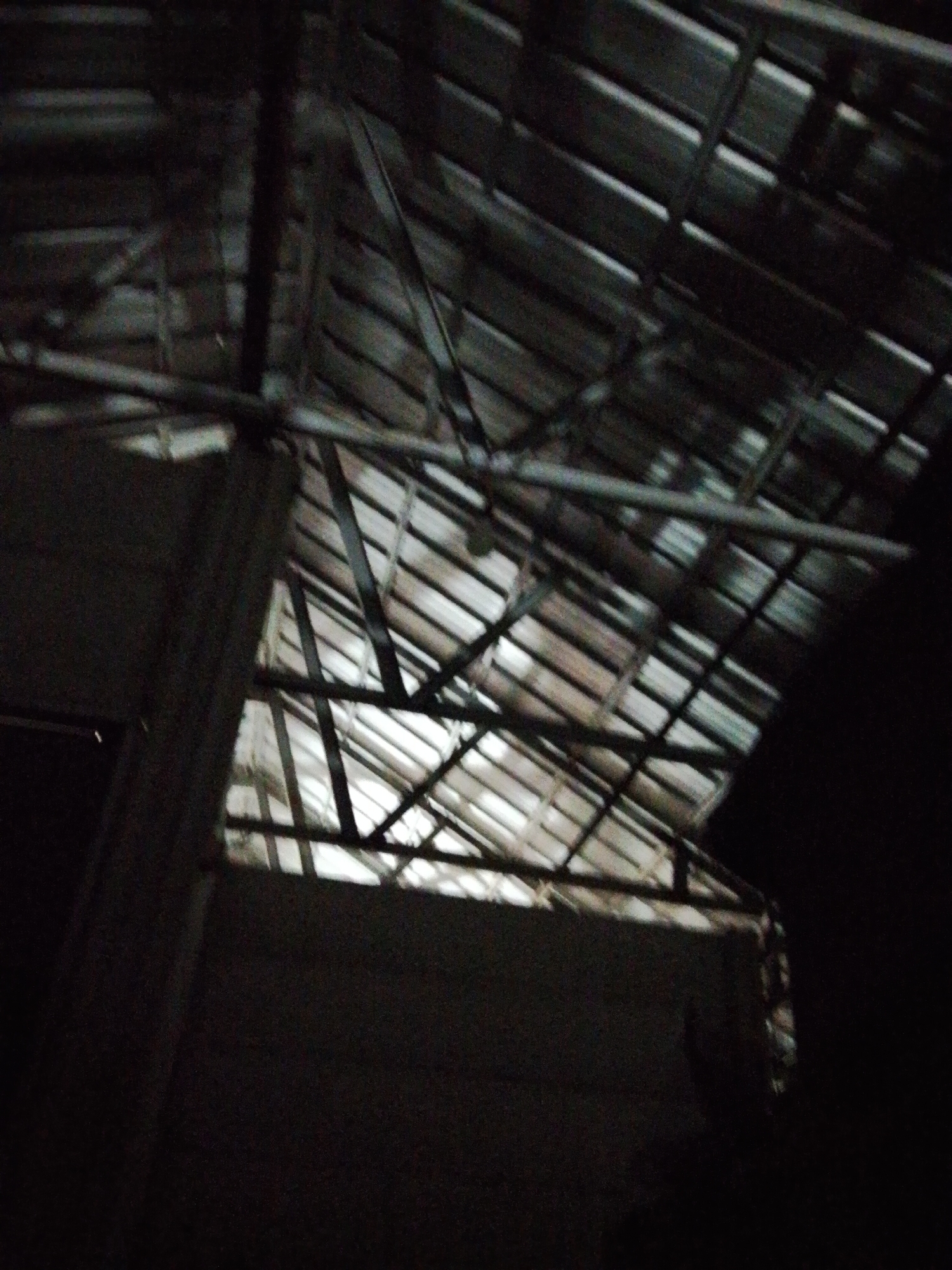 The image shows the interior of a building, possibly in Bukit Ketok, Indonesia, with a corrugated metal roof structure. The roof is supported by wooden or metal beams and rafters. A section of the roof is missing or damaged, allowing light to enter the building. The walls appear to be constructed of dark material, and the overall lighting is poor, with large areas in shadow.