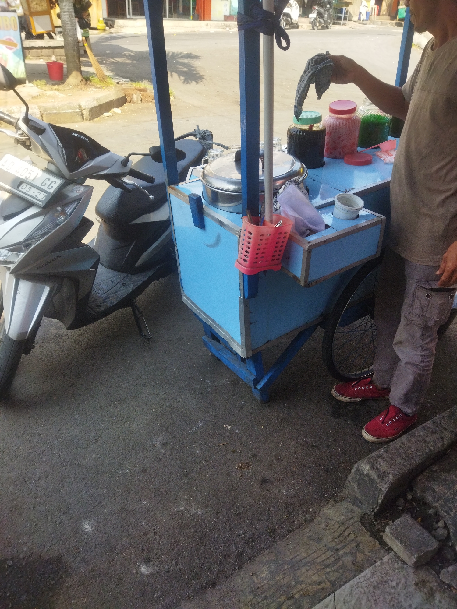 A street vendor is preparing drinks at a cart, likely in the city of Wonogiri, Indonesia. The cart is a light blue color and has a metal frame. It holds several glass jars filled with colorful liquids, including what appears to be syrup or juice. There are also metal containers and a red plastic utensil holder. The vendor, wearing a light-colored t-shirt and grey pants, is holding a cloth, possibly to wipe down the cart or containers. A grey Honda scooter is parked next to the cart. The ground is asphalt, and there's a concrete curb visible on the right side of the image. The overall scene suggests a typical street-side business operation in an Indonesian city.