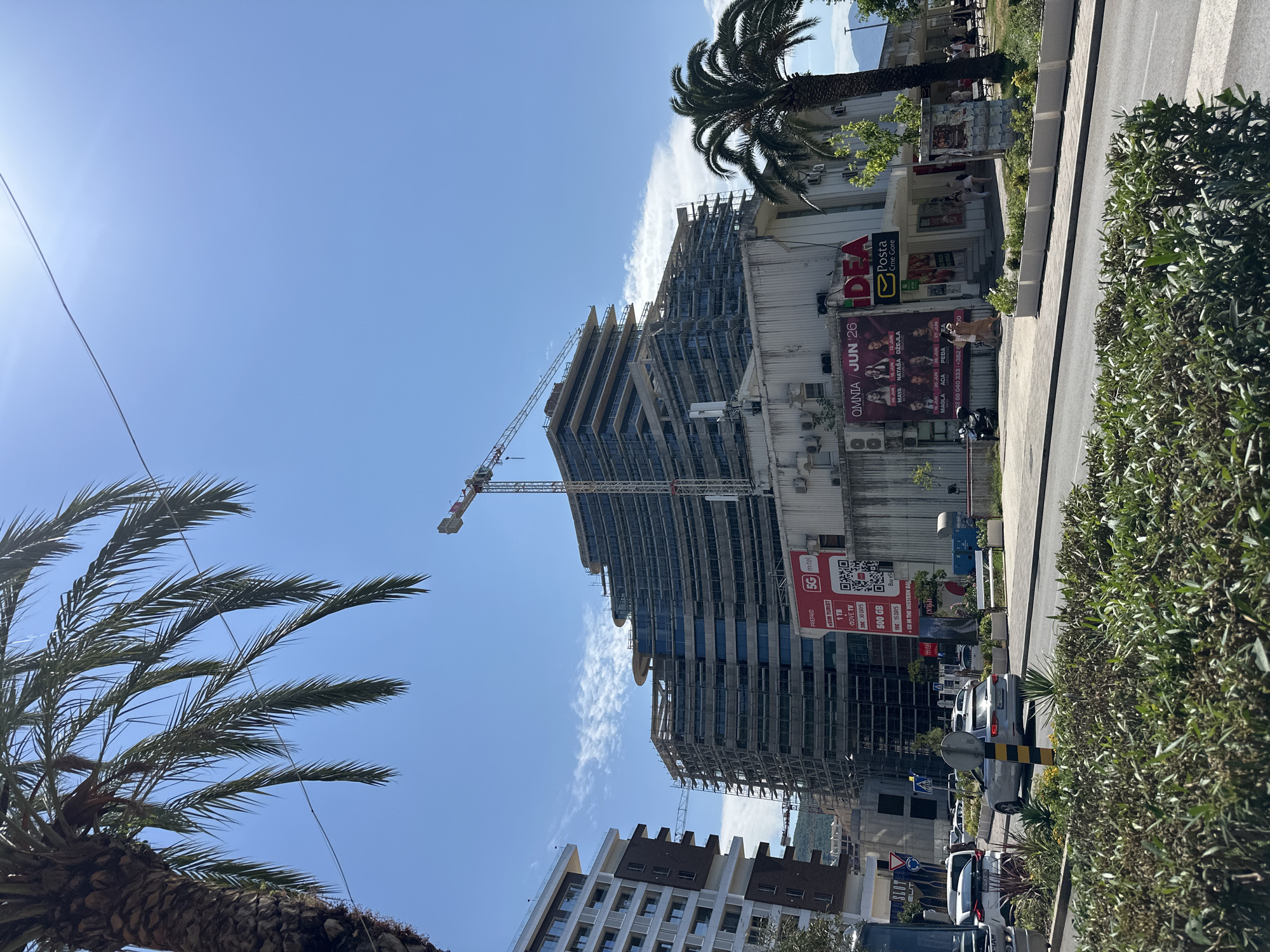 The image depicts a construction site in Budva, Montenegro, featuring a large, multi-story building under construction, encased in scaffolding, with a crane towering over it. To the left, a completed modern apartment building stands. In the foreground, a street with vehicles and lush green bushes suggests an urban setting. A palm tree, characteristic of the coastal region, is visible on the right. The sky is clear blue with scattered clouds, and the sun is bright, indicating a sunny day. Advertisements are visible on the lower portion of the construction site, including one for a mobile network operator and another for an event.