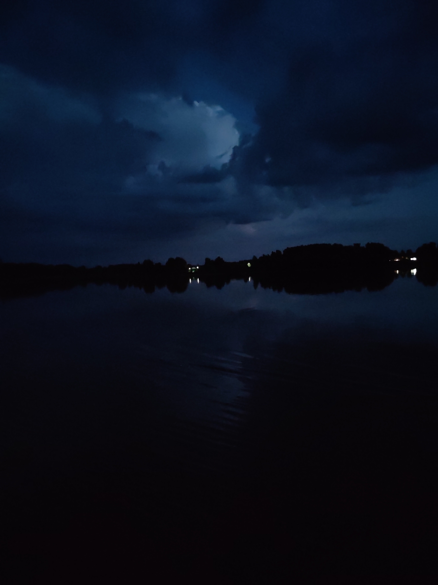 The image depicts a dark, moody scene of a body of water at night. The sky is filled with heavy, dark clouds, suggesting inclement weather, possibly a storm or impending rain. A faint glow of lights is visible along the distant shoreline, hinting at human habitation or a small settlement. These lights are reflected in the calm surface of the water, creating a soft, elongated shimmer. The overall impression is one of stillness and perhaps a touch of foreboding, amplified by the darkness and the brooding sky. The time of day is clearly night, and the weather appears to be overcast and potentially stormy. There are no visible people or specific objects that stand out, with the focus being on the natural elements of sky, water, and land. The location cues, such as the distant lights and the silhouette of the trees on the shore, are subtle but suggest a rural or semi-rural setting. No text is visible in the image.