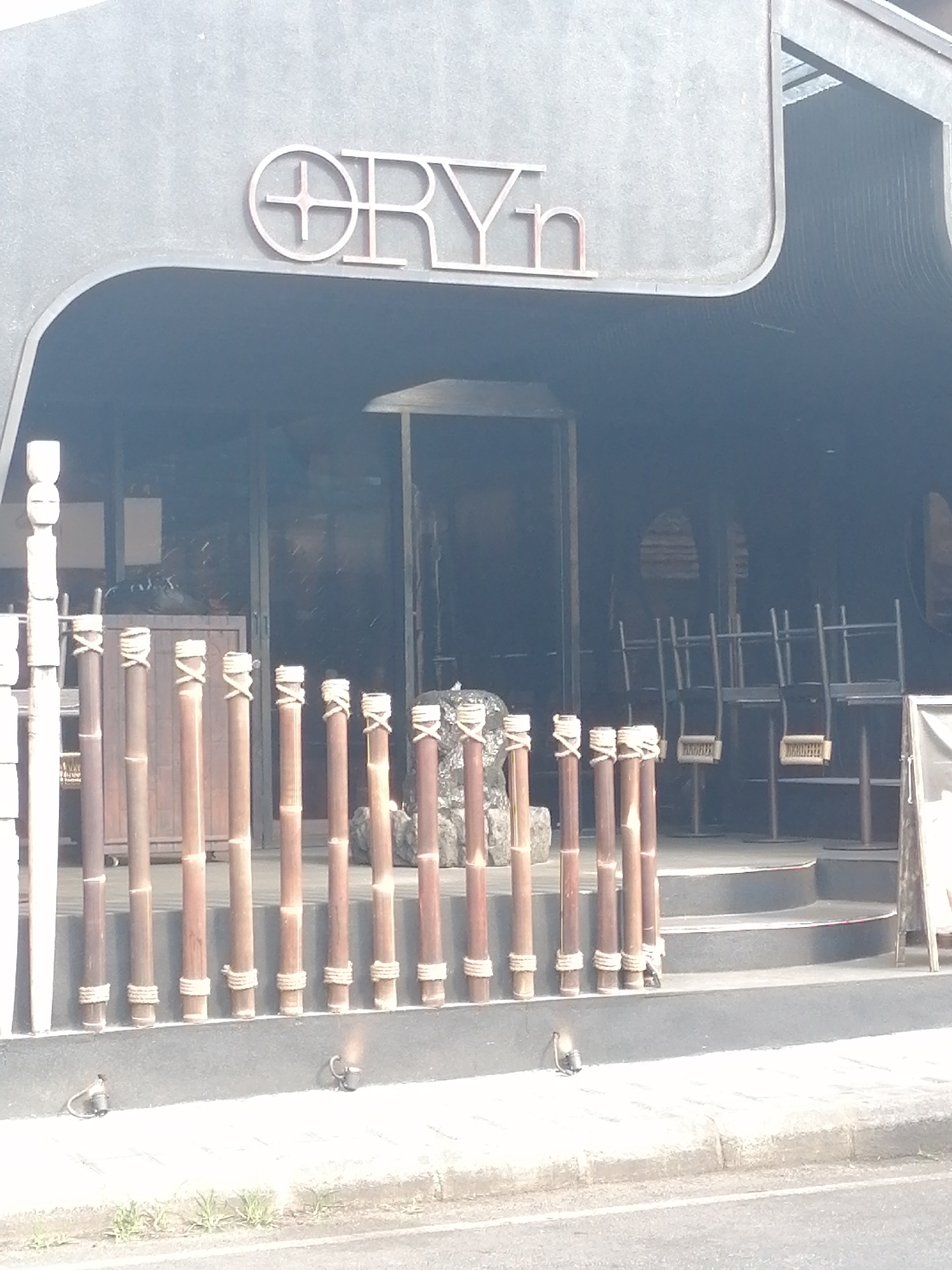 The image shows the exterior of a building with the sign "ORYn" in large, metallic letters. The building has a modern, dark gray facade with curved elements. A short bamboo fence with rope accents separates the entrance from the sidewalk. Behind the fence, steps lead up to a darkened doorway. Inside, chairs and tables are visible, suggesting an indoor seating area. The lighting is bright, indicating daytime. The environment appears to be urban, with a street visible in the foreground. The location is identified as Ubud District, Indonesia.