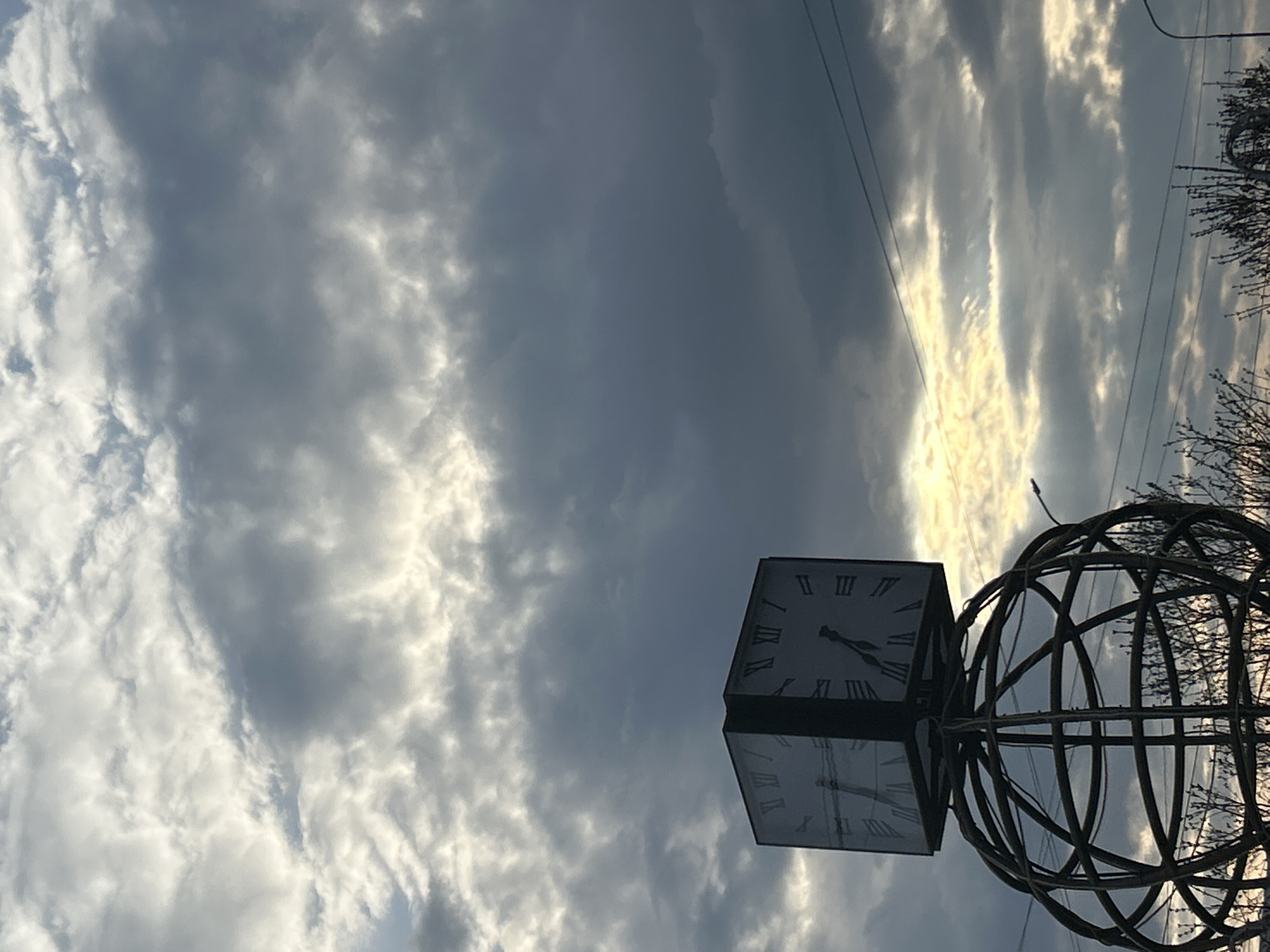 The image shows a clock on a metal globe structure in Chaikovsky, Russia. The sky is overcast with gray and white clouds. Power lines are visible in the upper right corner. Bare tree branches are also present on the right. The clock face features Roman numerals.