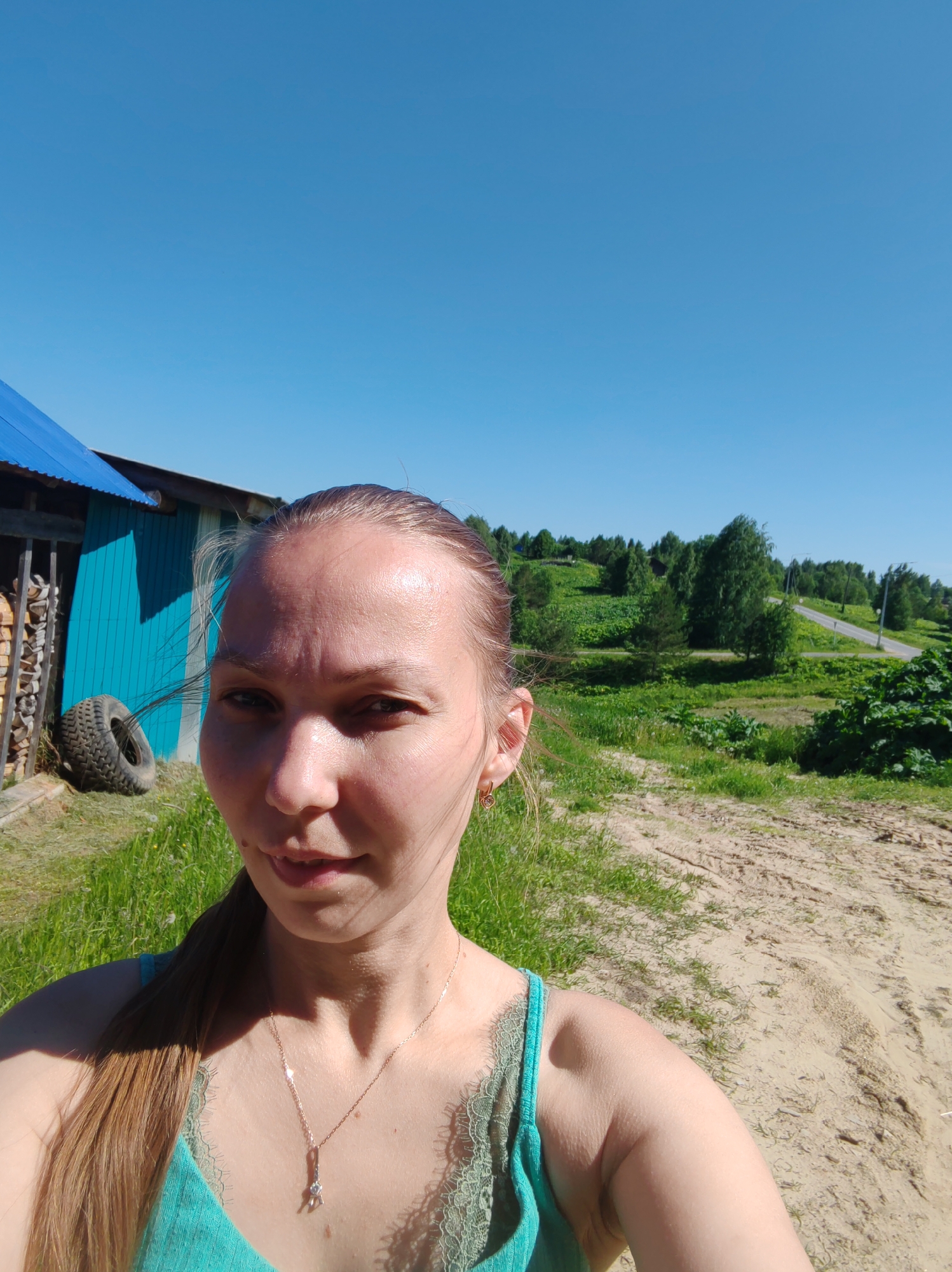 This is a medium-shot selfie of a woman in Chukhlem, Russia. She is outdoors on a sunny day, with a clear blue sky above. The woman has light brown hair pulled back in a ponytail and is wearing a turquoise tank top with lace trim and a delicate silver necklace. Her face is turned towards the camera, and she has a slight smile.
In the background, to the left, is a small blue building with a corrugated metal roof. Next to the building, there is a stack of firewood and a large black tire. The ground in front of the building is covered in green grass. To the right of the woman, a dirt road curves into the distance, flanked by lush green fields and scattered trees. A paved road is visible further back, winding through a hilly landscape. The overall impression is of a rural, summery setting.