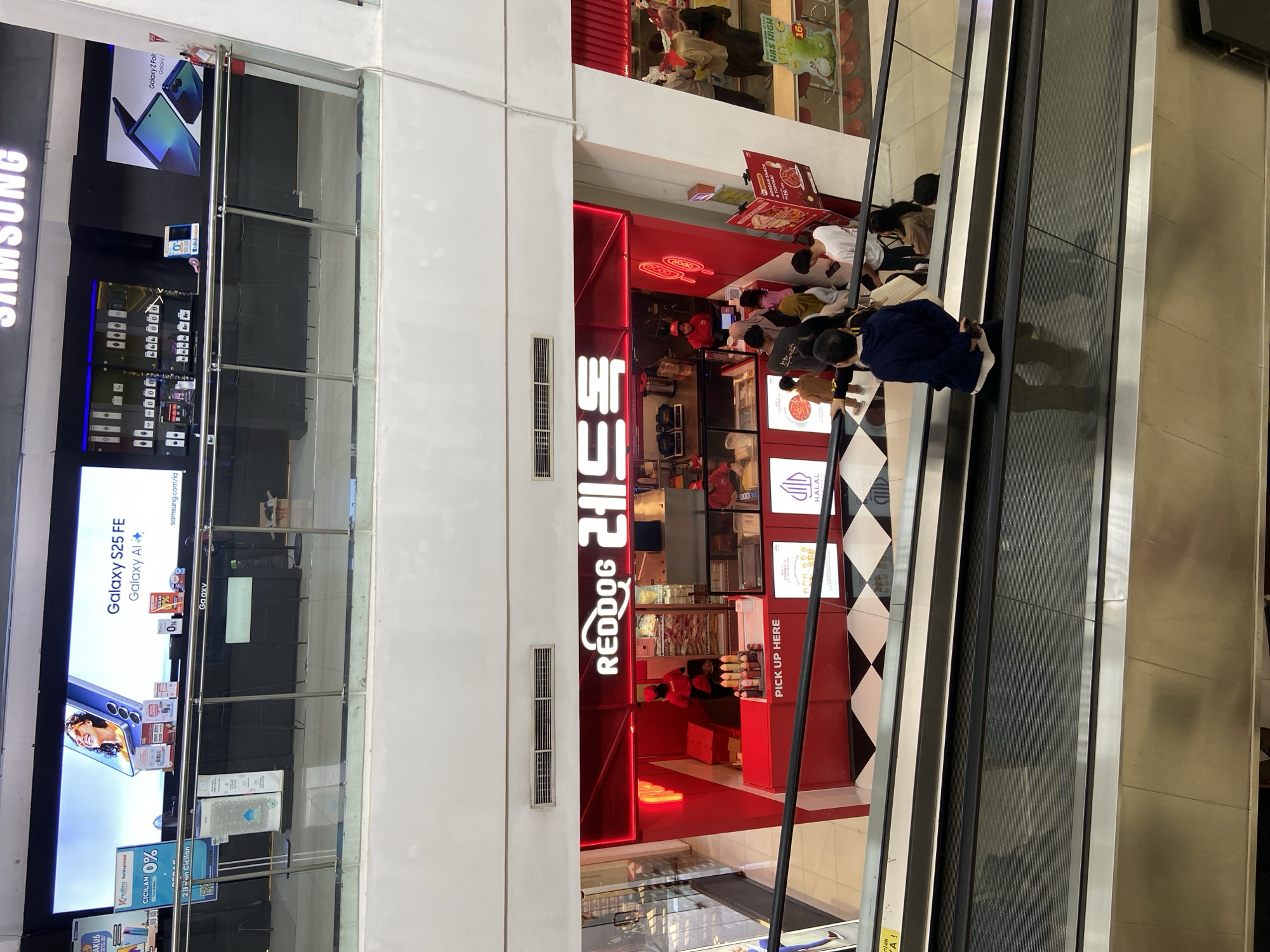In the city of Kudus, Indonesia, a top-down view captures an escalator in a shopping mall. The escalator is moving upwards, with several individuals riding it. Directly ahead and slightly to the right, a brightly lit red kiosk with the signage "REDDOG" and Korean characters is visible. The kiosk appears to be a food stall, with people queuing to order or pick up their food. Behind the kiosk, a display refrigerator filled with food items and a counter area are discernible. The overall environment suggests a busy commercial space, likely within a mall.