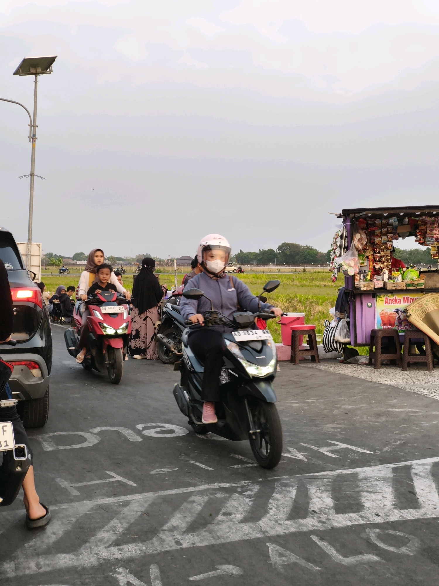 The image depicts a street scene in Desa Ngesrep, Indonesia, likely during daylight given the bright sky. Several people are present, primarily riding motorcycles. In the foreground, a person wearing a pink helmet and a mask rides a black scooter, with their face partially visible. To their left, another scooter carries a woman wearing a hijab and a young boy, both looking forward. Behind them, a woman in a black hijab stands near a street vendor's stall.
The street vendor's stall is purple and laden with various snacks and drinks. Several brown stools are placed in front of it. In the background, a solar-powered street lamp is visible on the left side of the frame. Beyond the road, a field of crops, possibly rice paddies, stretches out, bordered by trees and buildings in the distance. The road itself has white painted markings, including some that appear to be words or symbols that are partially obscured by shadows. The overall atmosphere suggests a bustling local area, possibly a market or a busy thoroughfare.