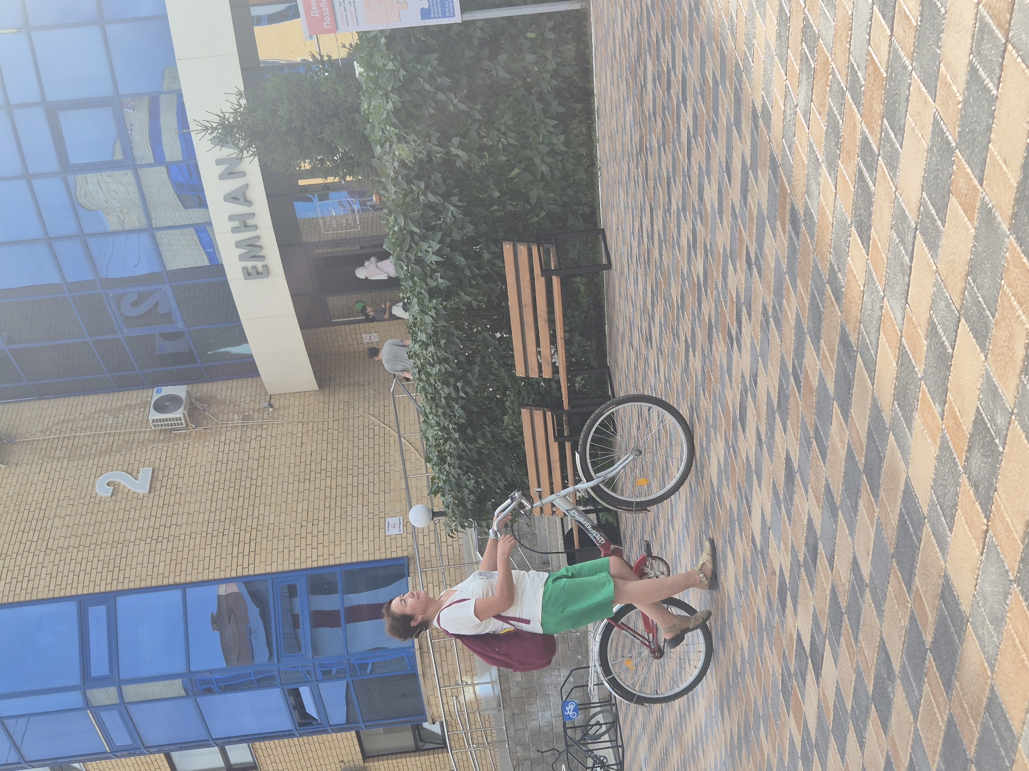 A middle-aged woman with dark hair pulled back walks her bicycle on a paved area in front of a building with a brick facade and blue-tinted windows. The woman wears a white t-shirt, green shorts, and beige sandals, and carries a red backpack. Her bicycle is silver with red accents. The building features the word "EMHANAV" above an entrance, and the number "2" is visible on the brickwork. To the right of the building is a dense green hedge, and behind it, a small evergreen tree is visible. A wooden bench with a black metal frame sits in front of the hedge. The paving stones are arranged in a diamond pattern of beige, gray, and brown. The scene appears to be outdoors during the daytime, with the sun casting a slight glare on the building's windows. The location is identified as being in Kokshetau, Kazakhstan.