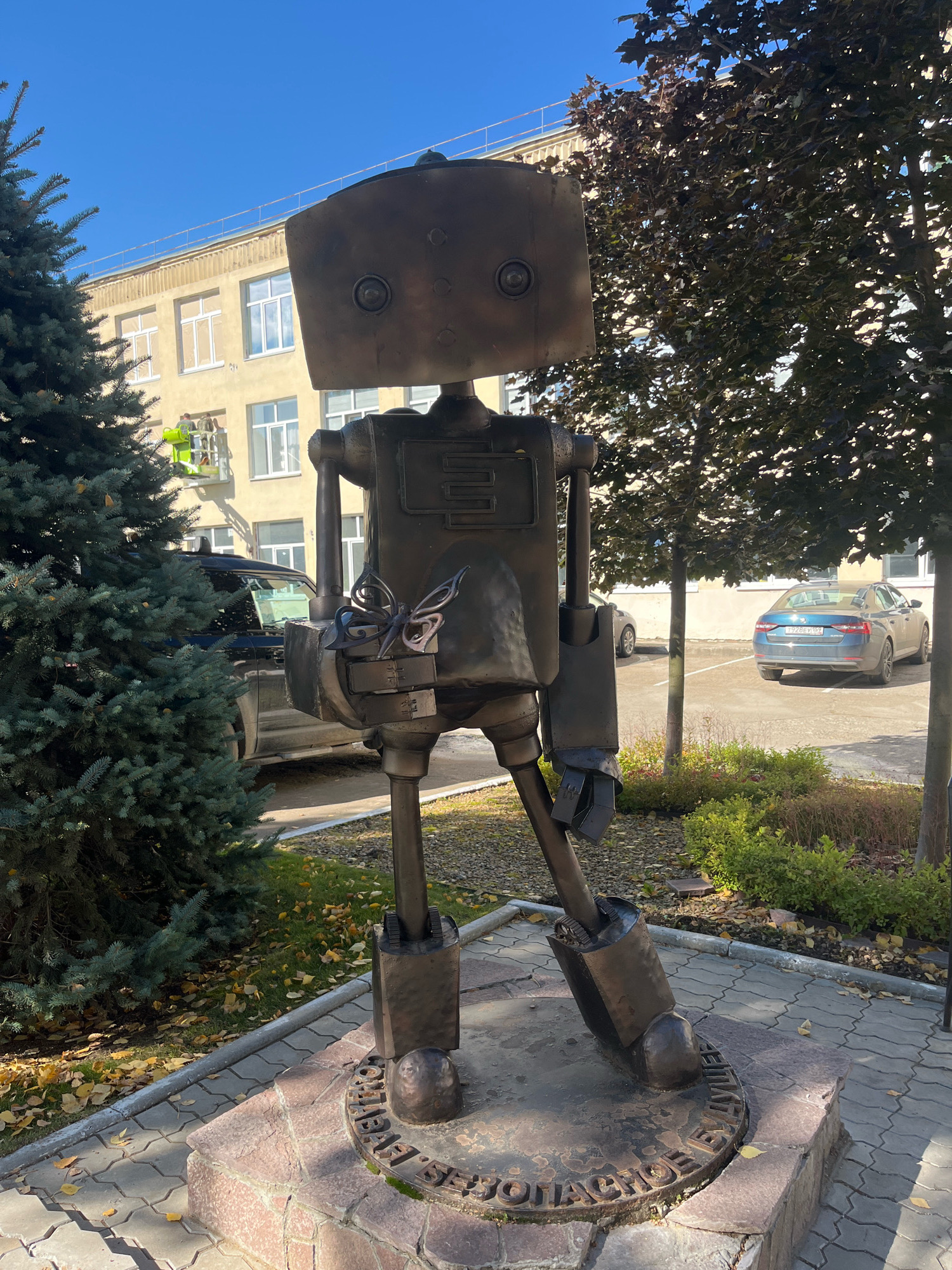 A bronze statue of a robot stands on a stone pedestal on a sidewalk in Chaikovsky, Russia. The robot is constructed from scrap metal, with a rectangular head, circular eyes, and a metallic body. A small, intricate metal butterfly is attached to its chest. Behind the robot, a light yellow building with white-framed windows is visible. Two workers in a green cherry picker are cleaning the facade of the building. To the left of the robot, a tall, dark green evergreen tree stands. To the right, trees with dark foliage and some fallen autumn leaves on the ground are present. A blue car is parked in the background. The sky is clear and blue. The statue is located in front of the Chaikovsky State Theater and Concert Hall.