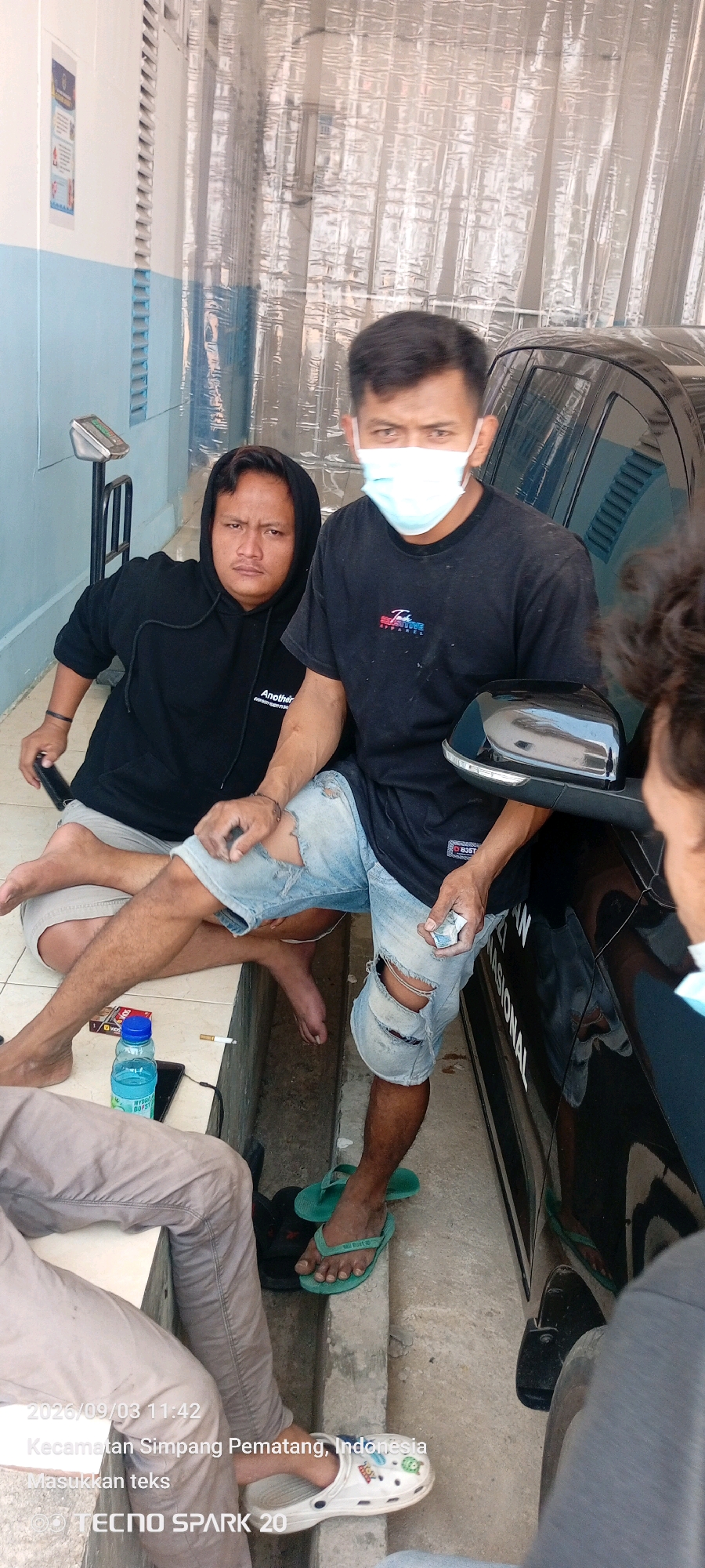 Three individuals are present in an outdoor, possibly industrial or service area. On the left, a person wearing a black hooded sweatshirt and light-colored shorts is seated, looking towards the camera. In the center, another individual, wearing a black t-shirt and distressed denim shorts, is also seated and looking at the camera; this person has a light blue face mask on. To the right, partially visible, is a third person, also wearing a black t-shirt. A dark-colored car is parked beside the seated individuals. Various objects are scattered on the ground, including a plastic water bottle, a cigarette, a cell phone, and a digital scale can be seen on the left side of the image. The ground appears to be paved with concrete. The background consists of a wall with vertical blinds or louvers, covered by a translucent, textured material. Text overlays on the image indicate the date and time "2023/09/03 11:42" and the location "Kecamatan Simpang Pematang, Indonesia".