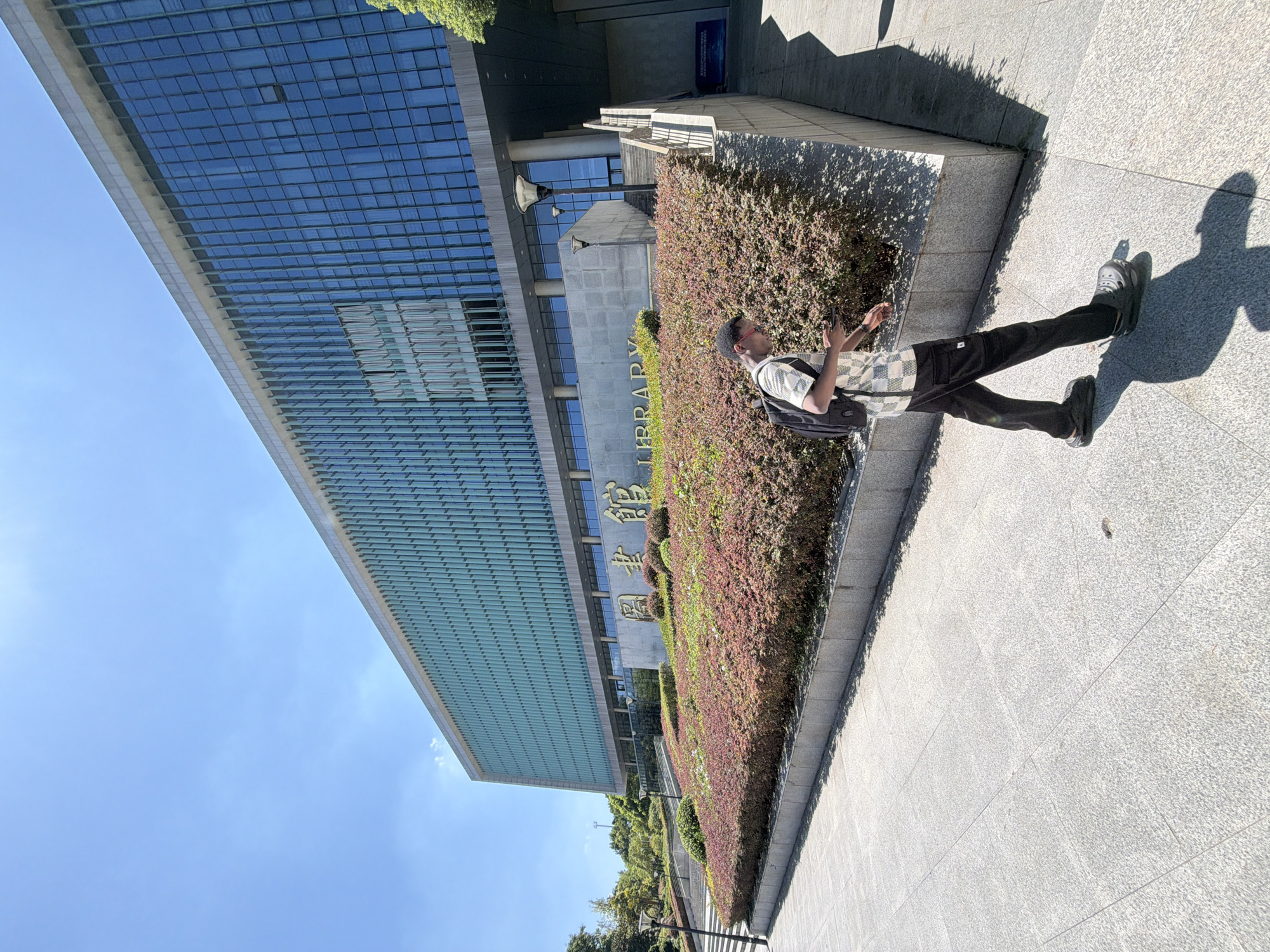 A man walks on a paved sidewalk in front of a large, modern library building in Jiangjin, China. The building has a glass facade with green vertical accents and a gray concrete base. A sign with gold lettering, partially obscured by foliage, reads "LIBRARY". The man is wearing a checkered shirt, black pants, a black backpack, and dark clogs. He is looking down at his phone as he walks. Lush, reddish-pink bushes line the path leading to the library entrance. The sky is clear and blue, and the sun casts long shadows on the ground.
