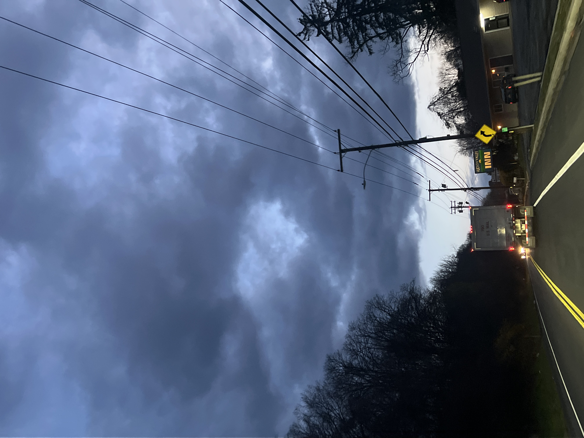 The image captures a nighttime scene on a road in what appears to be the City of Newburgh, United States of America. Dominating the left side of the frame is a dramatic, cloudy sky, hinting at possible inclement weather. Power lines stretch across the upper portion of the image, leading towards utility poles along the roadside. On the right, a two-lane road with double yellow lines is visible, with a truck trailing behind. The sign for the Hudson Valley Inn is also visible. The scene evokes a sense of dusk or twilight.