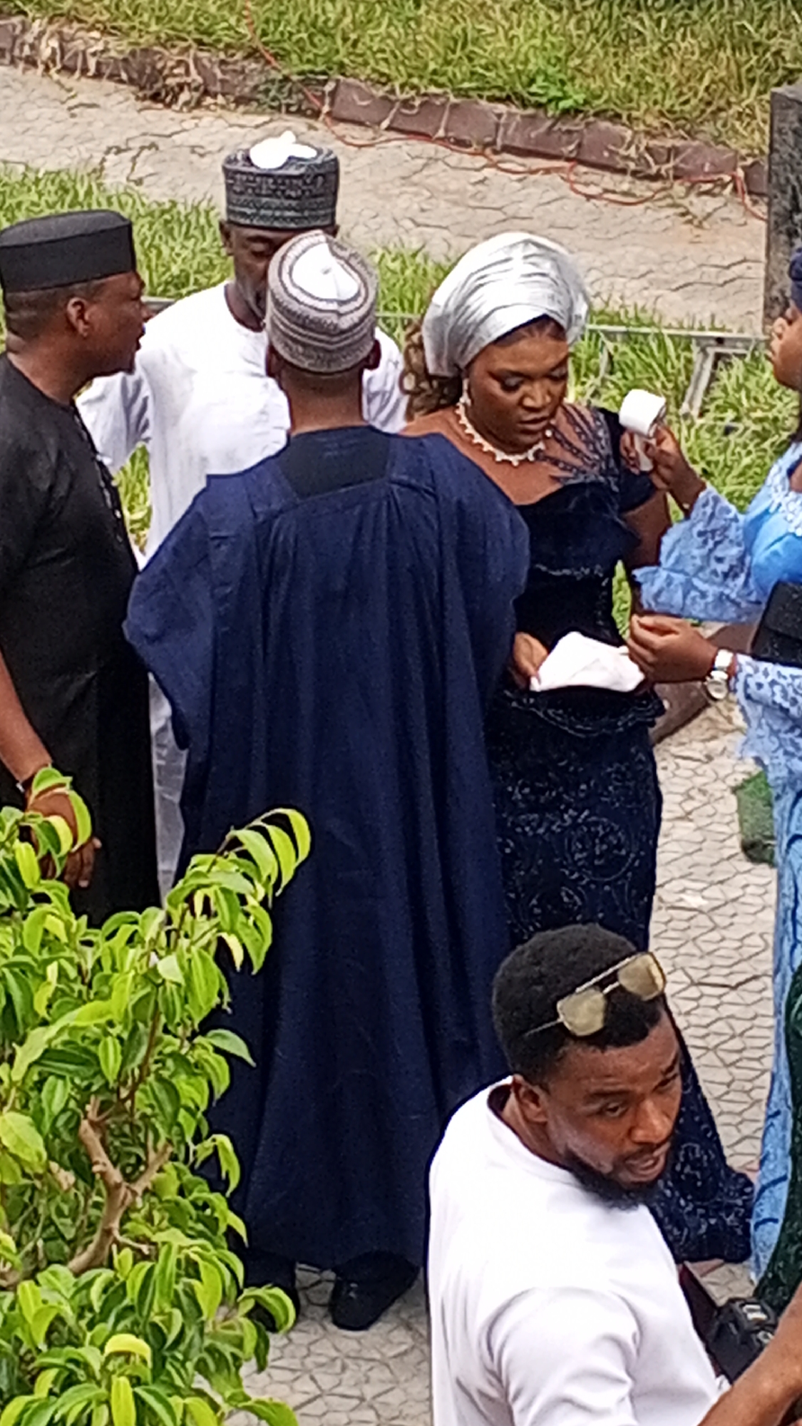 In Lokoja, Nigeria, a group of people are gathered outdoors on a paved area near greenery. Five people are partially visible in the foreground, while two more are in the background. One man wears a black cap and attire, and another is dressed in white. The person in the center wears a blue robe. A woman in a silver headwrap and dark, patterned dress is attended to by another woman in a blue dress. A man with sunglasses rests atop his head wears a white shirt. The setting suggests a social event or gathering in the city of Lokoja.