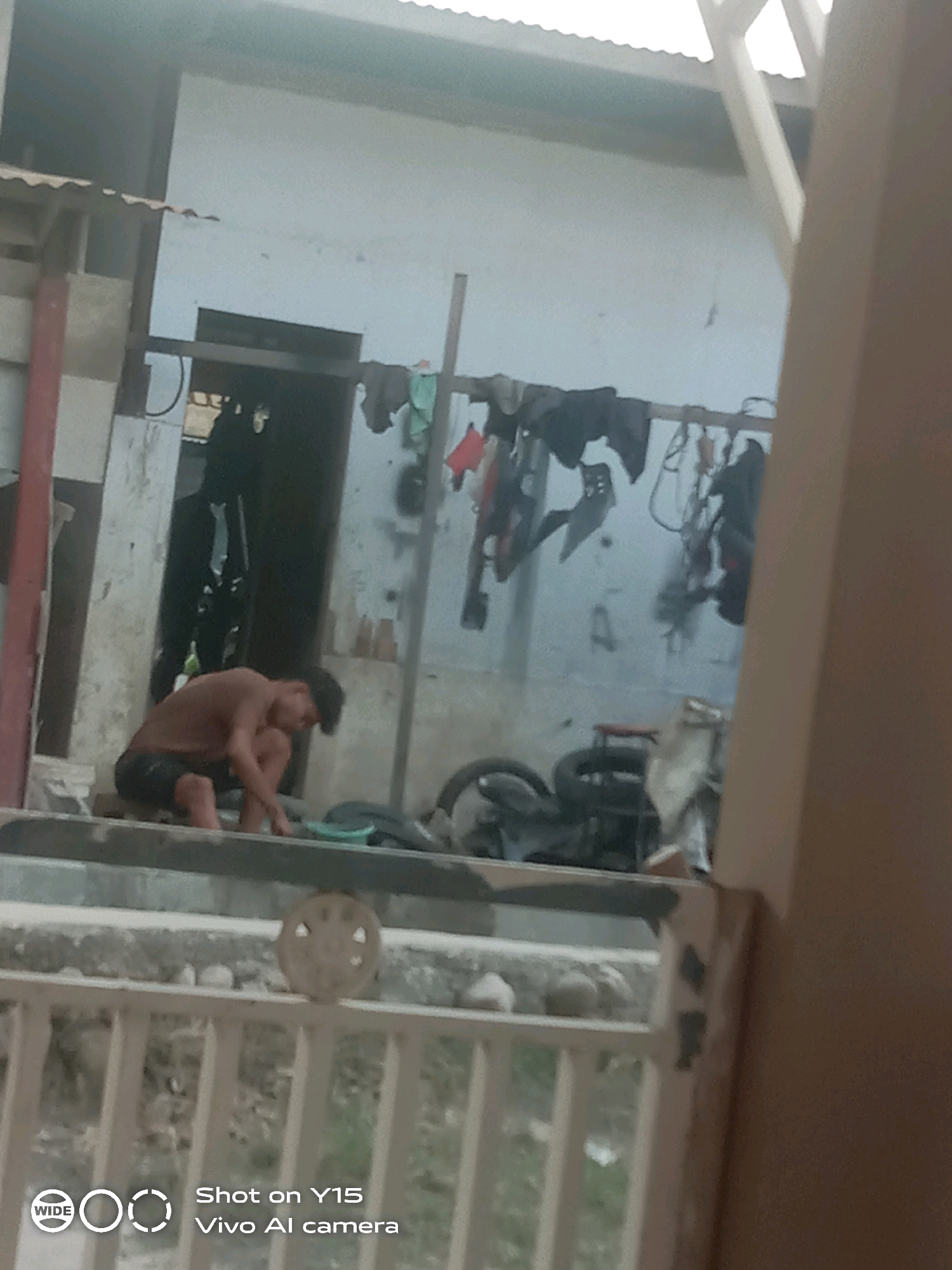 In Beurawang, Indonesia, a young person is seen squatting near a concrete structure, possibly washing something in a small green basin. They are wearing dark shorts and a short-sleeved shirt. Behind them, various items are hanging from a railing, including dark clothing and what appear to be motorcycle parts. A partially visible motorcycle is also present to the right of the young person. The scene appears to be an outdoor, utilitarian area, with a corrugated roof overhead and a white building in the background. The lower part of the image shows a white railing in the foreground. The overall atmosphere is that of an informal, possibly domestic, workspace.